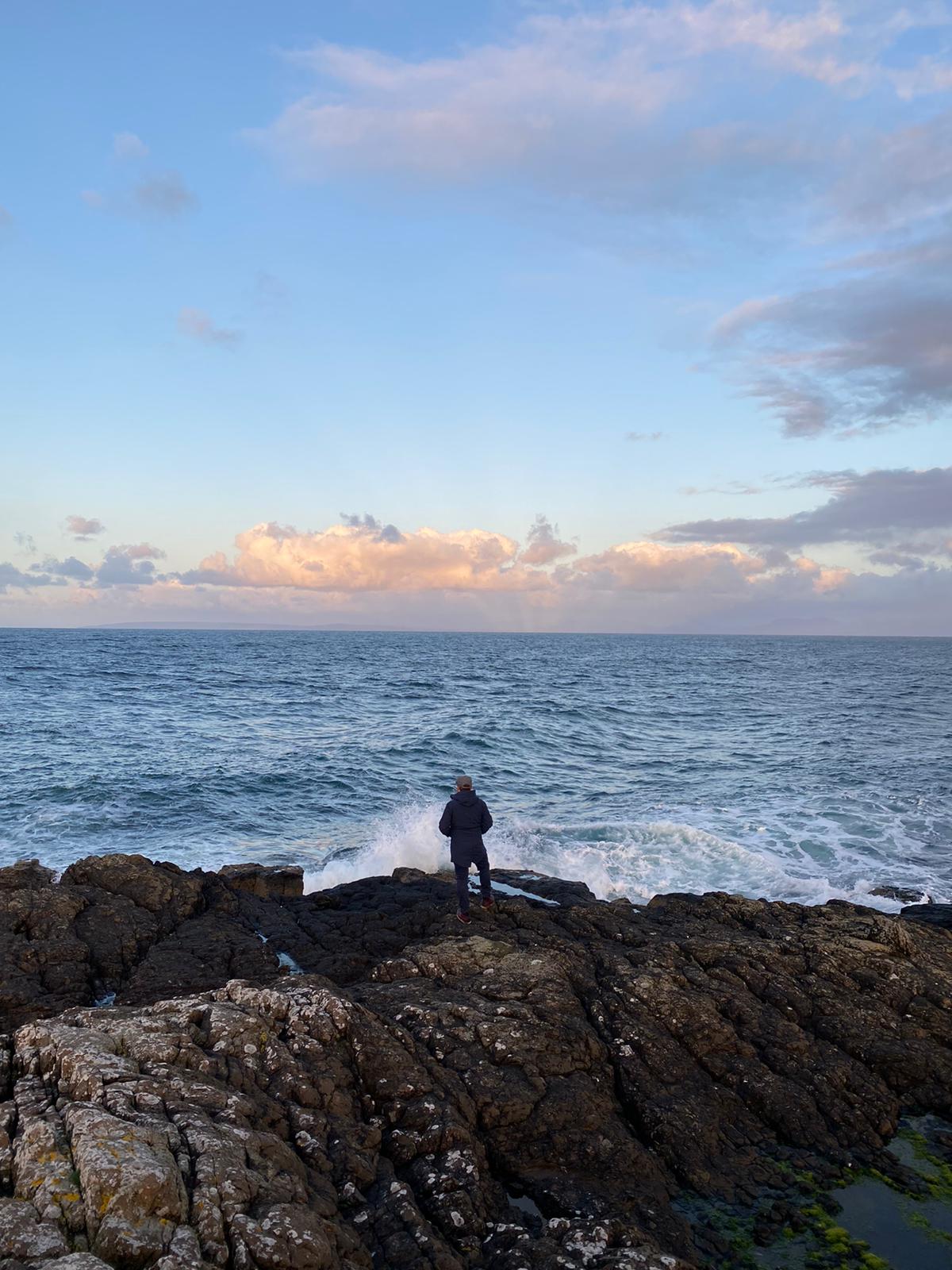 A person stands at the edge of a shoreline on rocks, while the sea crashes against the rocks. They look into the horizon.