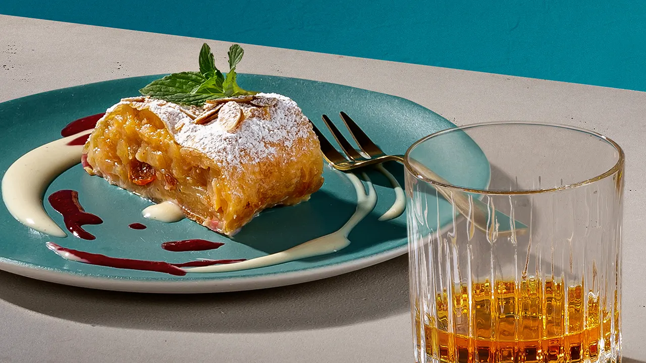A glass of whisky next to a plate of apple strudel.