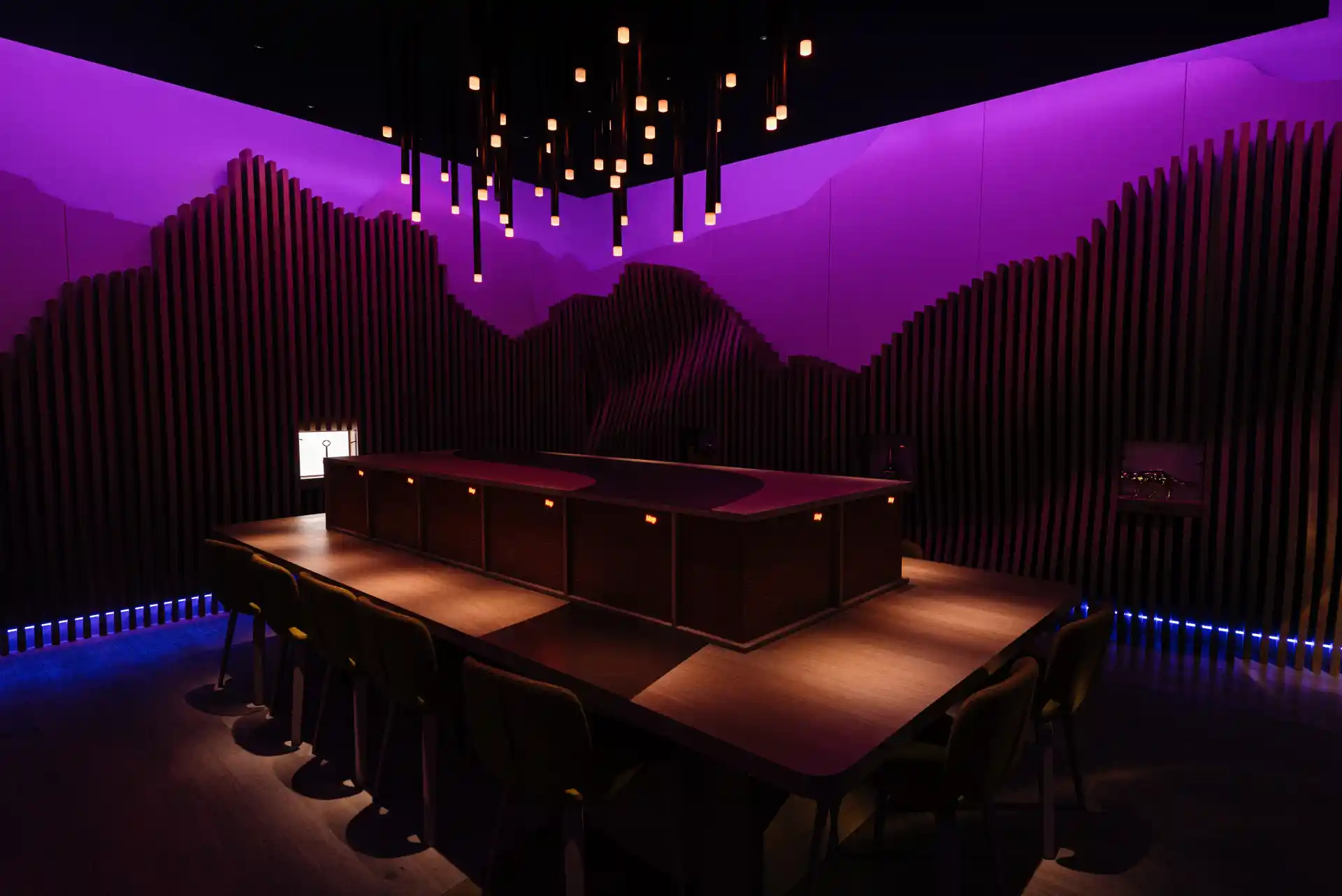 The interior of a whisky tasting room, wavy wooden panels on the walls and a large rectangular wooden table in the middle of the room. Several chairs and a large wooden box on the table. Purple wall and spotlight on the celling