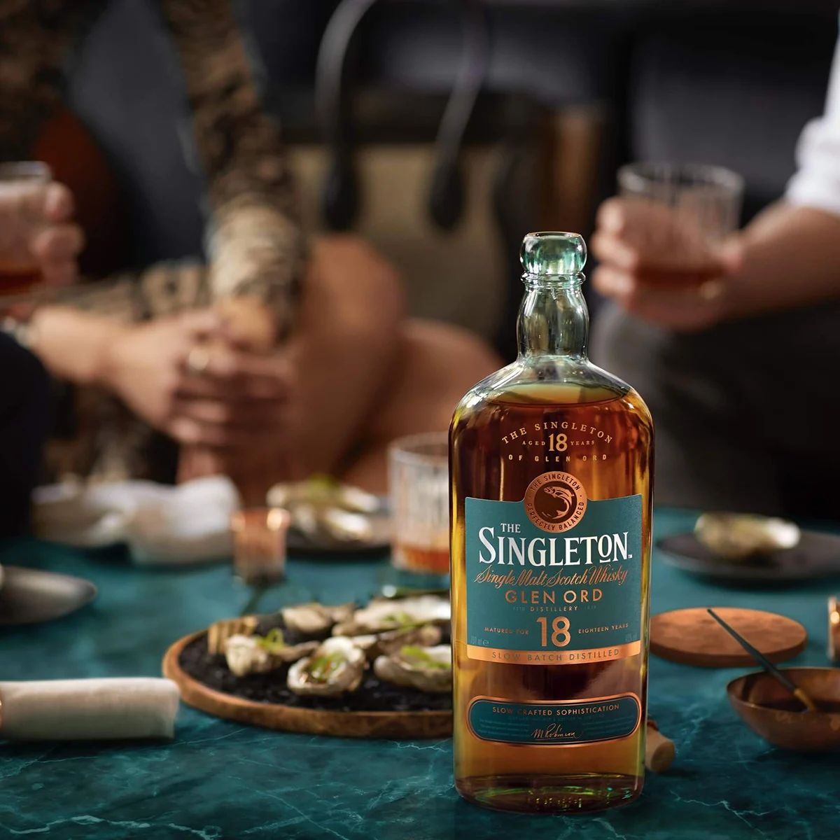The Singleton of Glen Ord 18 Year Old Single Malt Scotch Whisky Bottle on a table in front of a plate of oysters while a group of people gather to enjoy