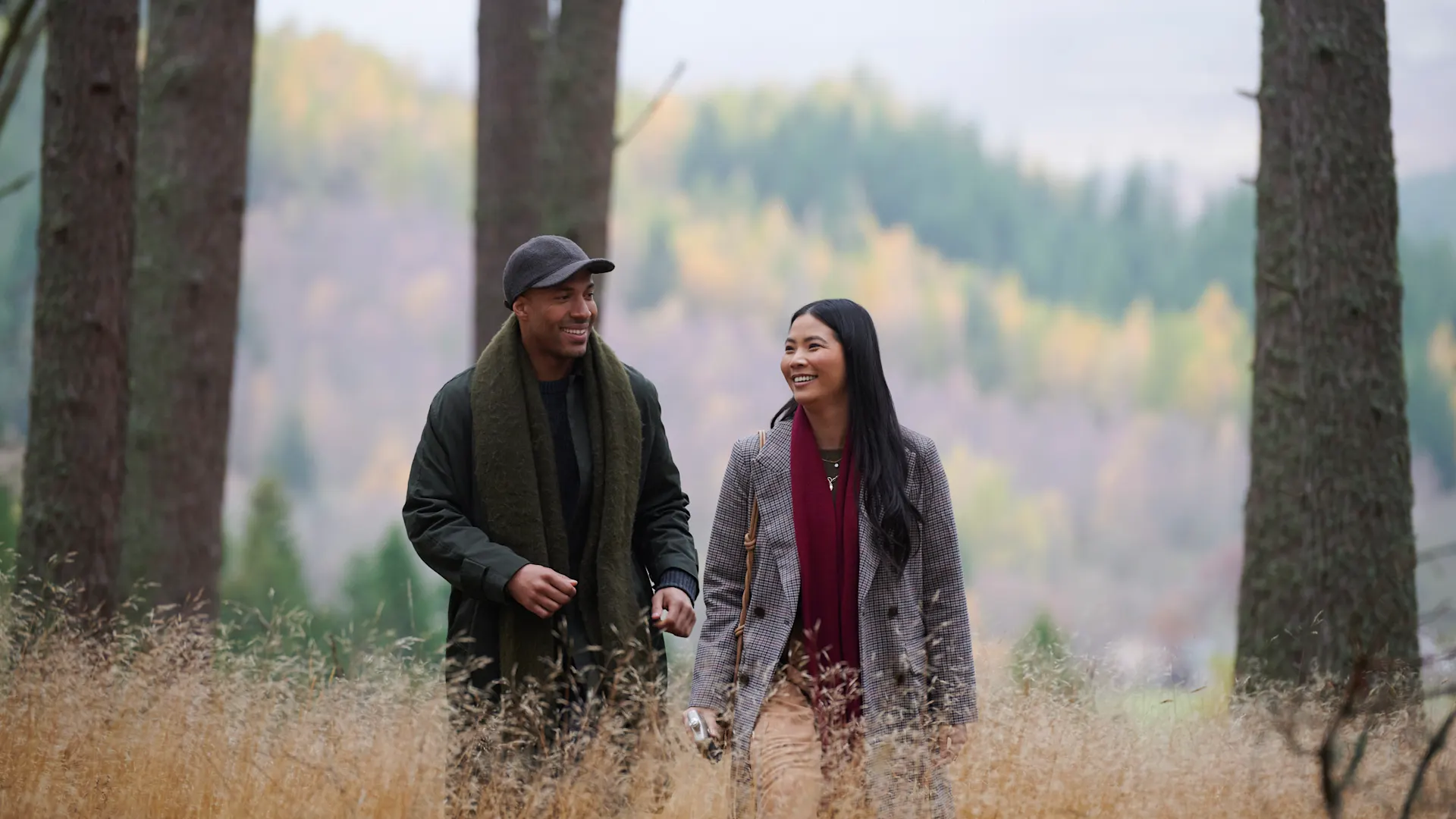 Een man en vrouw lopen door de Highlands in de herfst. Ze glimlachen en de vrouw houdt een heupfles vast.