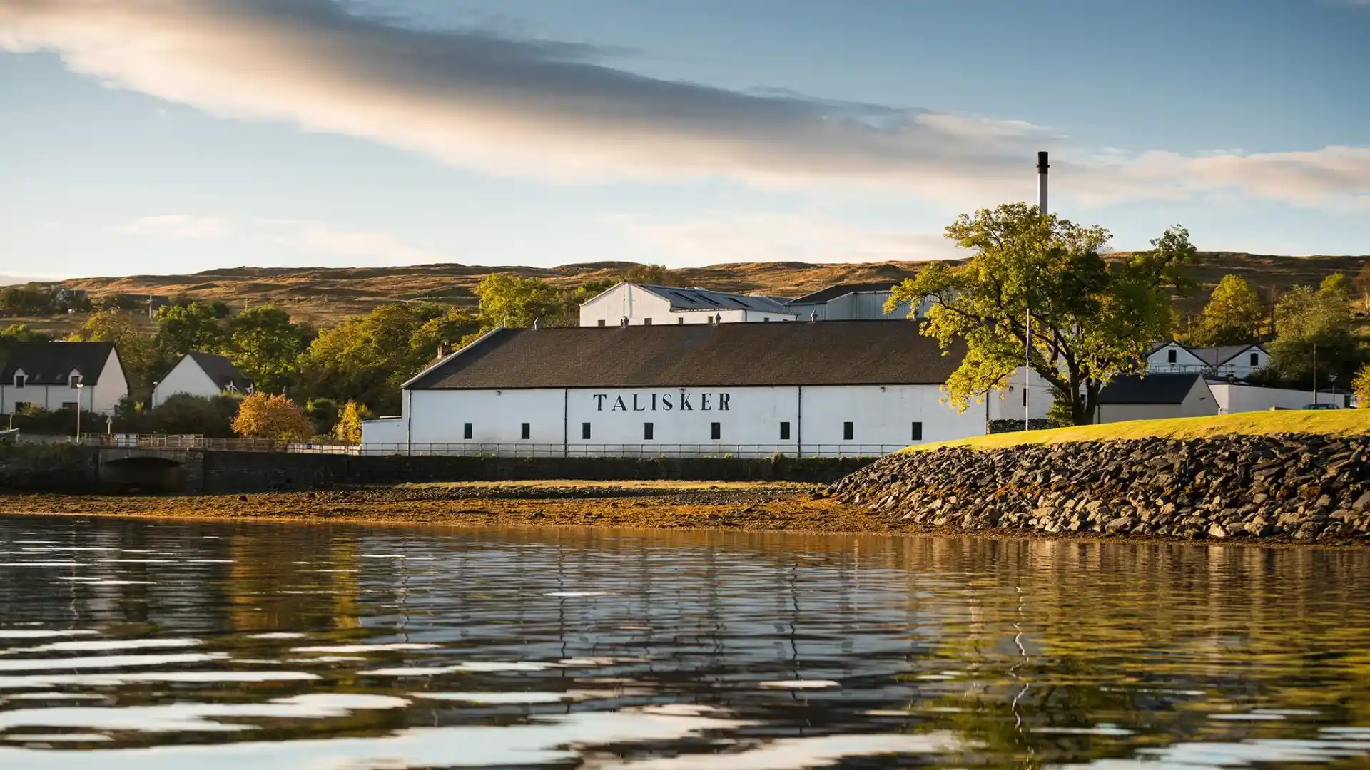 Het witte gebouw van de Talisker distilleerderij ligt aan een water met een kiezelstrand. Er zijn andere witte gebouwen in de buurt en groene heuvels erachter. De woorden 'Talisker' staan in grote zwarte letters op één gebouw.