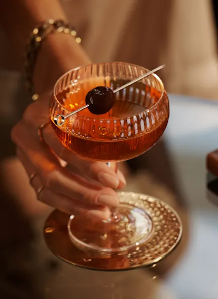 A woman holds the stem of a coupe glass containing a Manhattan cocktail