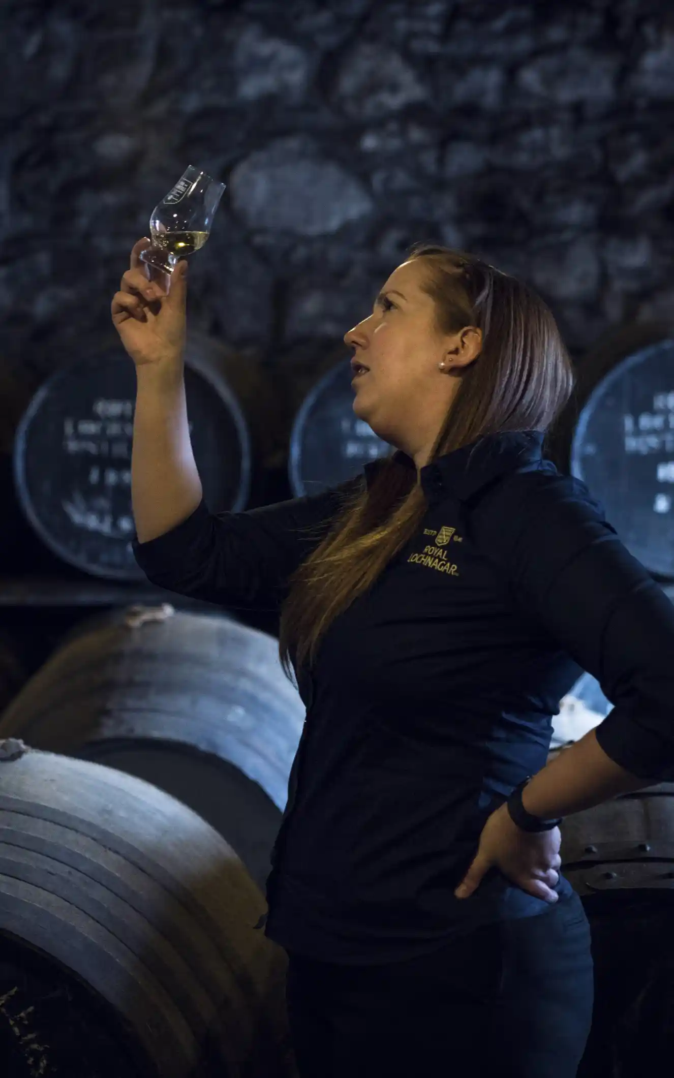 A woman holds up a dram glass containing whisky and inspects it, while standing in front of lots of wooden barrels.