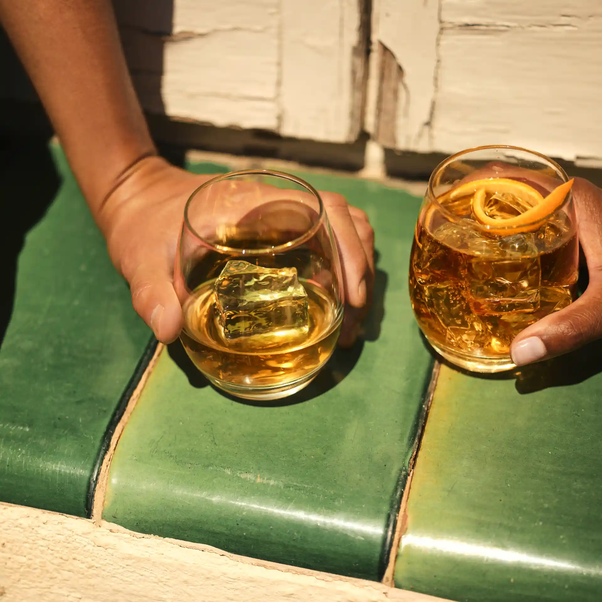 Two glasses of whisky sit on a green tile, both glasses are held by hands