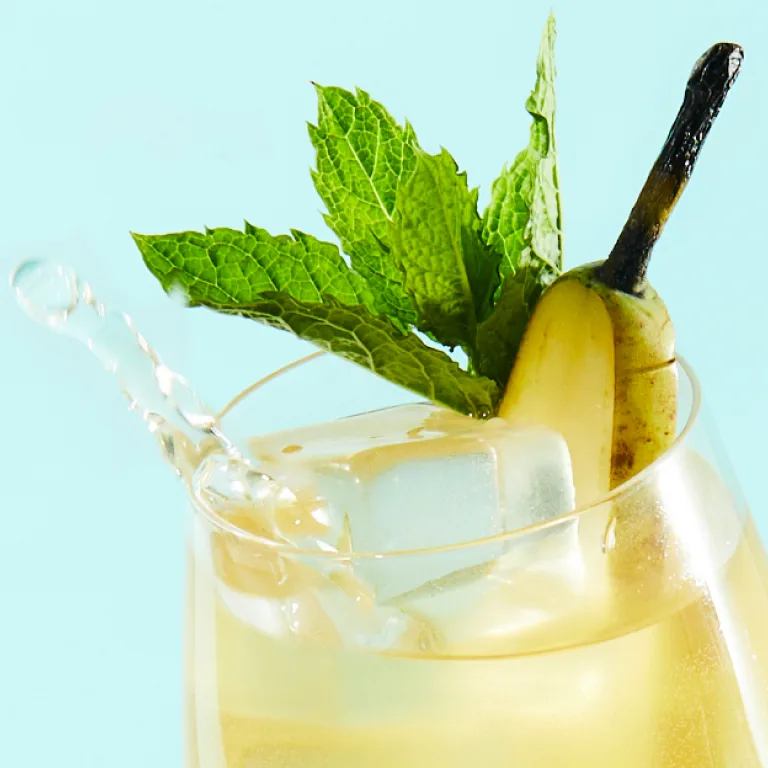 Een spetterend glas met gele vloeistof tegen een lichtblauwe achtergrond. Het glas is gevuld met ijs en gele vloeistof, gegarneerd met een munttakje en een schijfje peer.