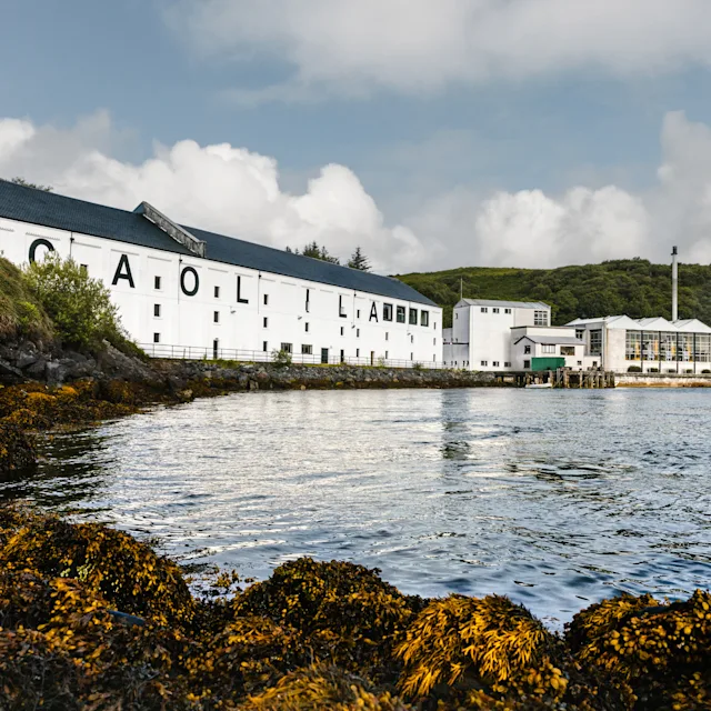 Learn more The white buildings of Caol Ila distillery sit on a calm body of water surrounded by green and yellow banks. On one part of a building, the words Caol Ila are printed in large black letters.