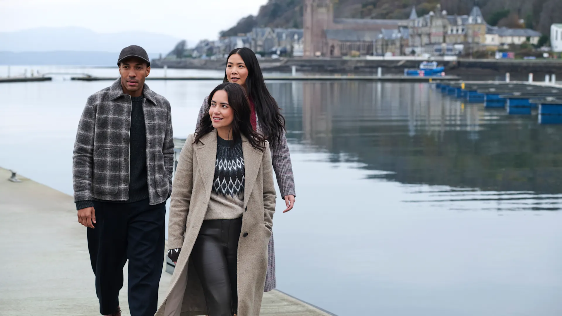 Drei Freunde gehen am Ufer der Bucht von Oban entlang. Hinter ihnen, gegenüber dem Meer, befinden sich Gebäude. 
