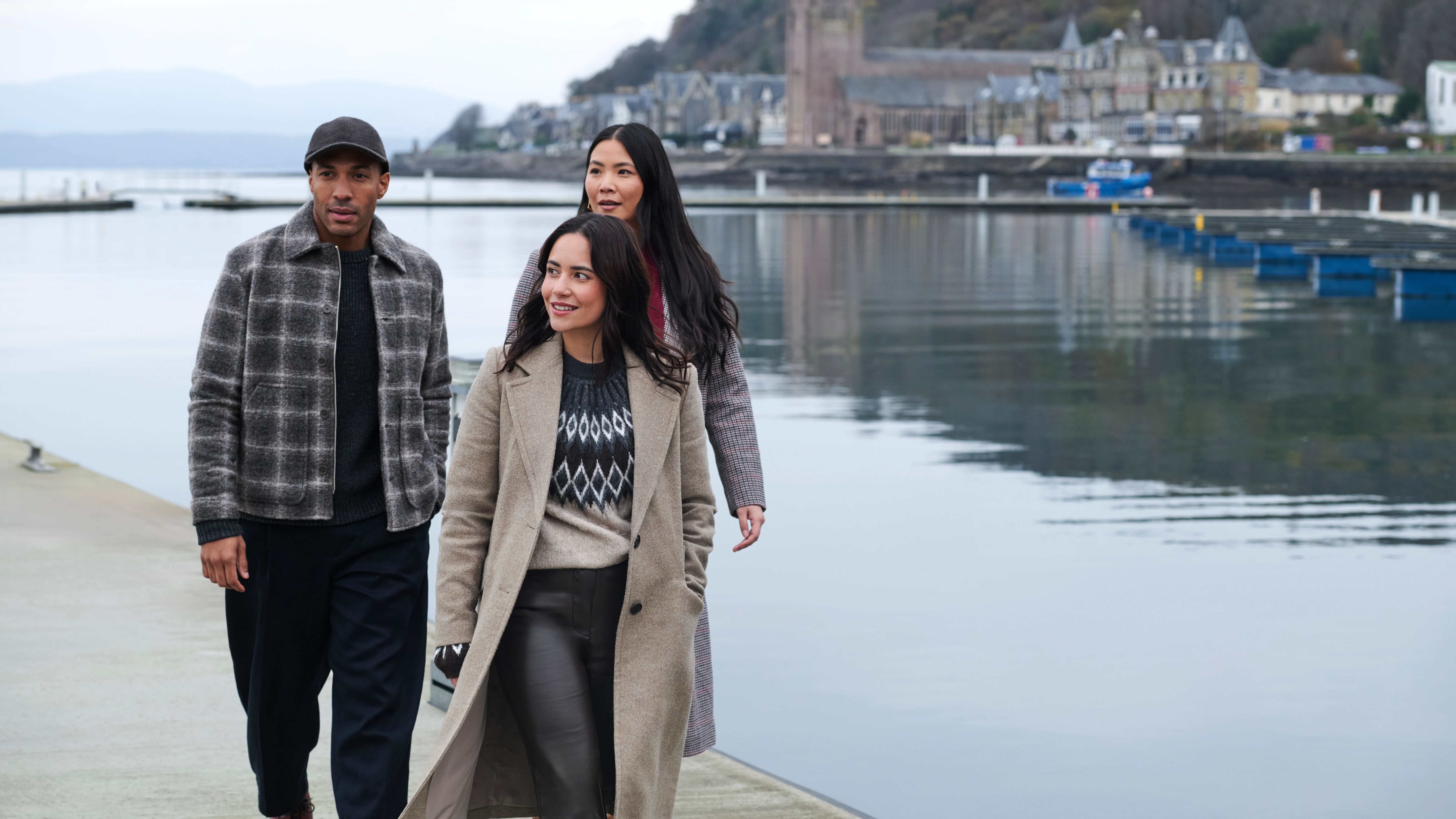 Three friends walk along the waterfront of Oban Bay. There are buildings behind them, across from the sea. 