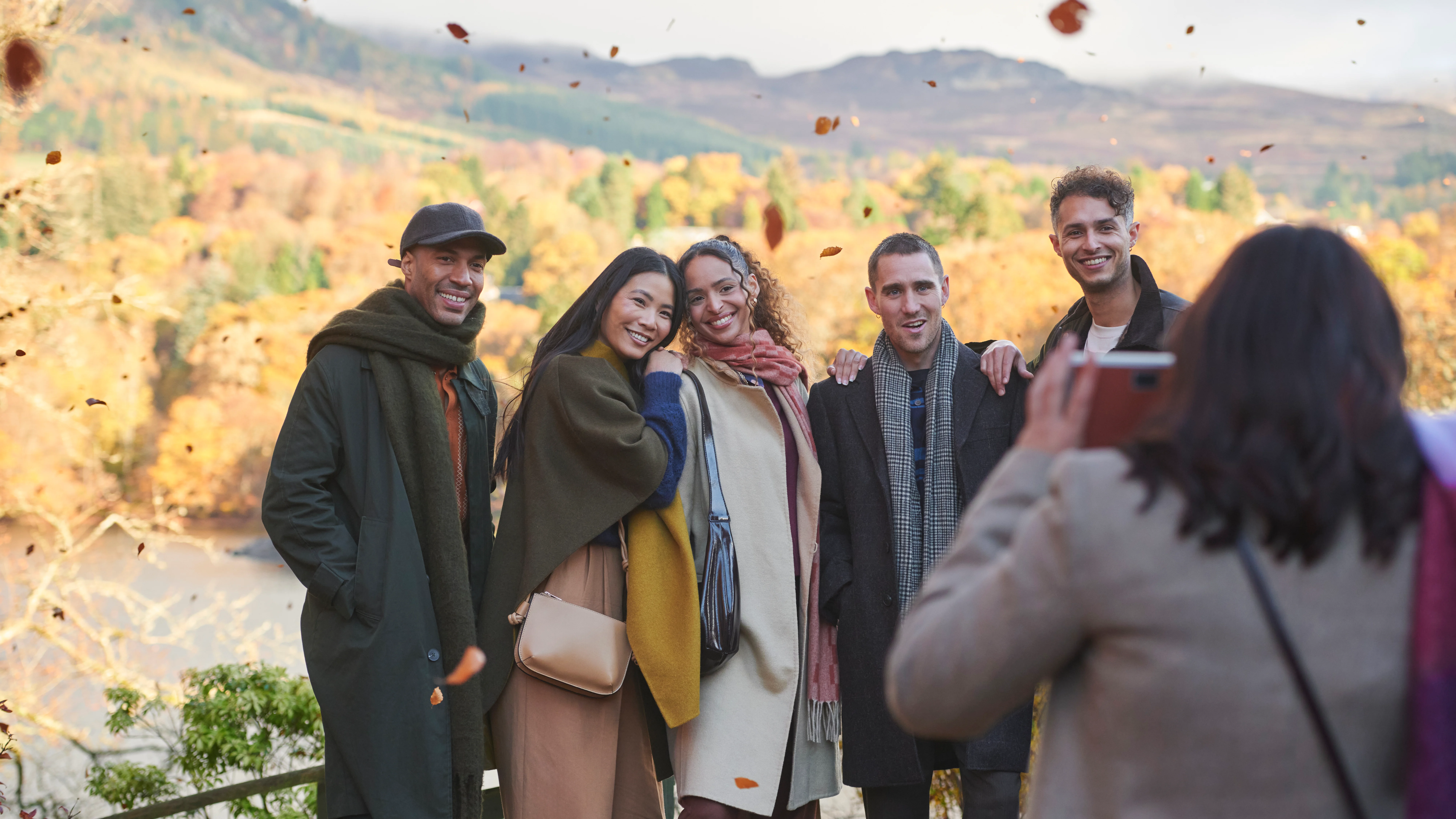 Eine Gruppe von Freunden steht zusammen und lächelt einem anderen Freund zu, der ein Foto von der Gruppe macht. Hinter der Gruppe liegt ein See und eine weitläufige schottische Landschaft. Sanft fallen die Blätter von den Bäumen um die Gruppe herum.