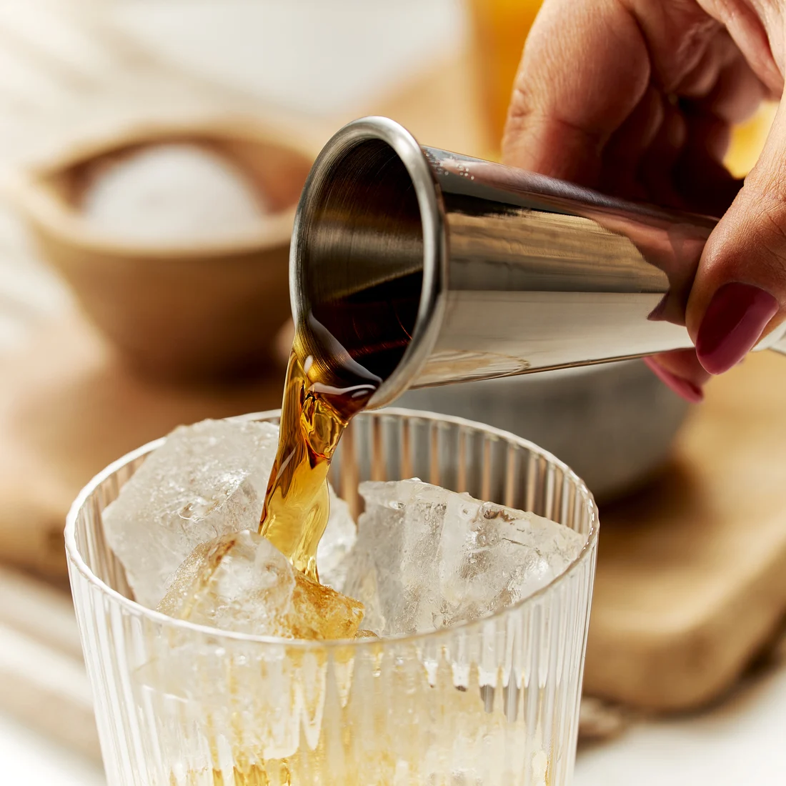 Een donkere gedistilleerde drank wordt uit een zilveren jigger in een met ijs gevuld glas gegoten.