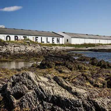 A rocky shore and still body of water sits in front of the white building of Port Ellen distillery, against a backdrop of a bright blue sky.