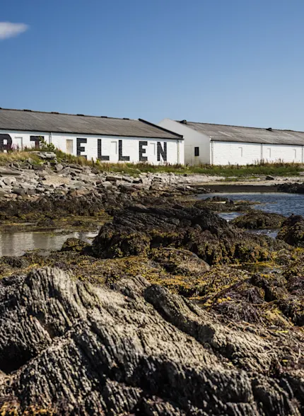 A rocky shore and still body of water sits in front of the white building of Port Ellen distillery, against a backdrop of a bright blue sky.