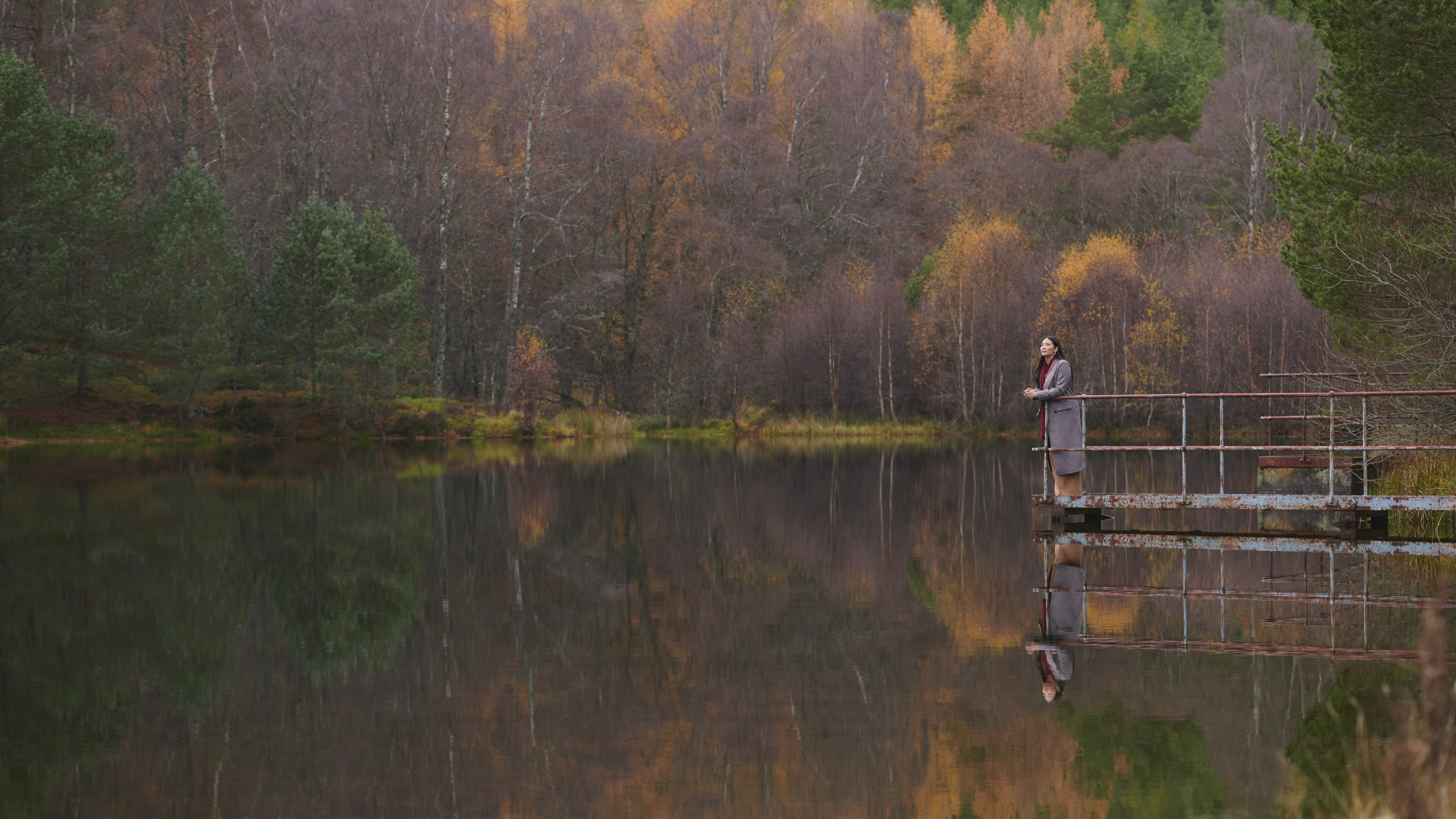 A woman stands on a metal jetty, looking out at a still loch. The loch is surrounded by autumnal trees which are reflected in the loch's water.