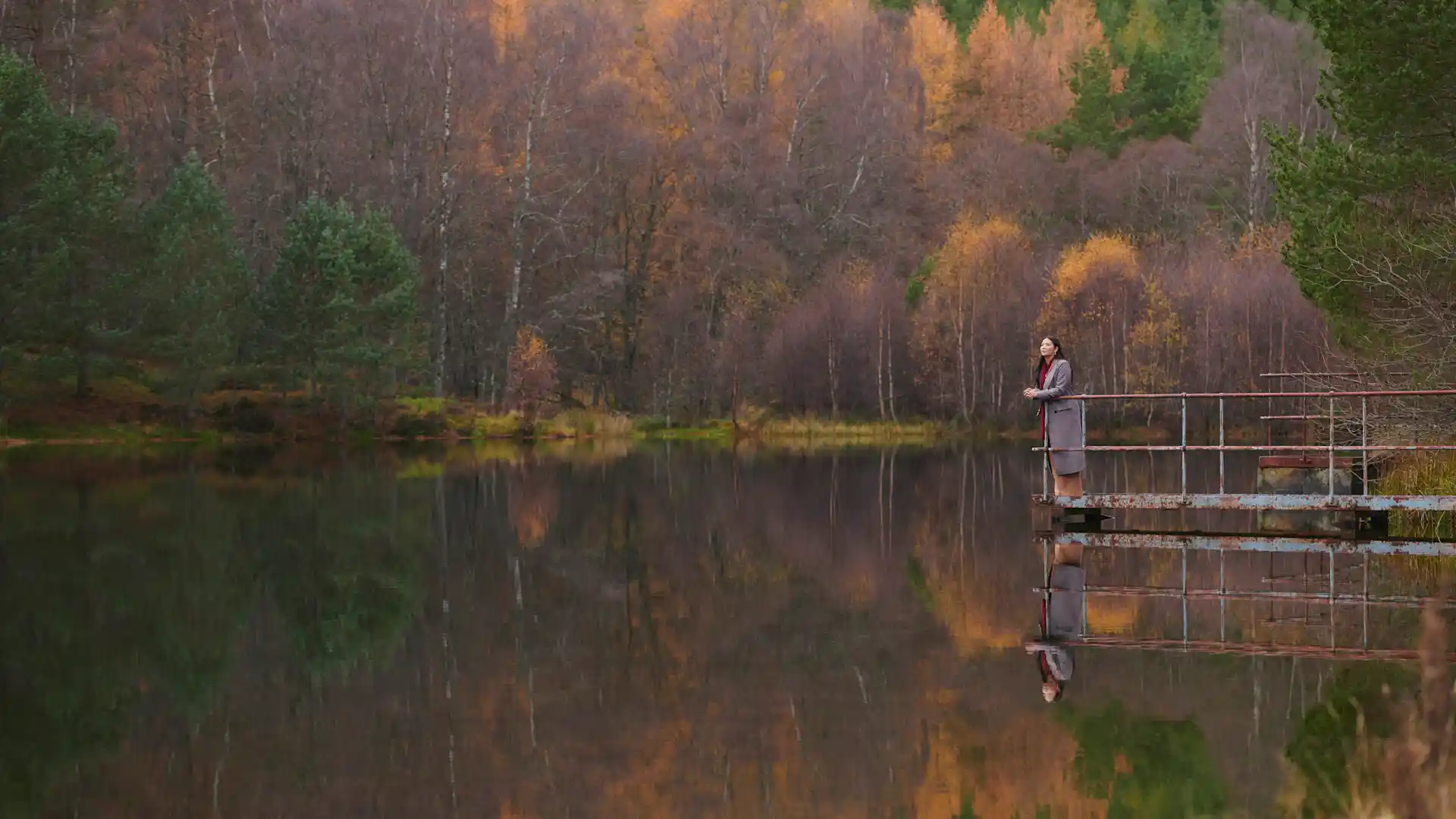 Een vrouw staat op een metalen steiger en kijkt uit op een stilstaand loch. Het loch wordt omringd door herfstbomen die weerspiegeld worden in het water van het loch.