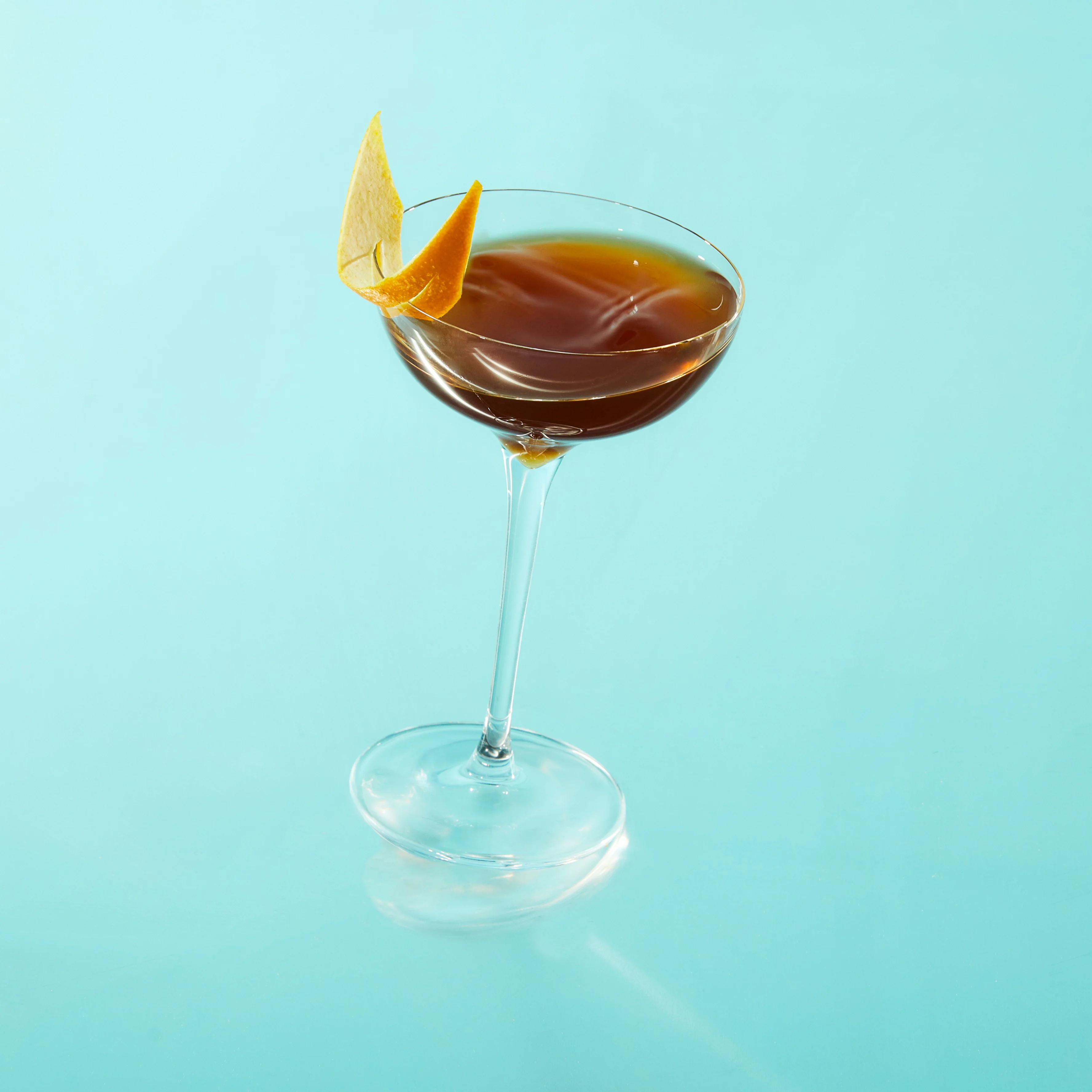 A cocktail in a coupe glass with an orange twist garnish against a blue background.