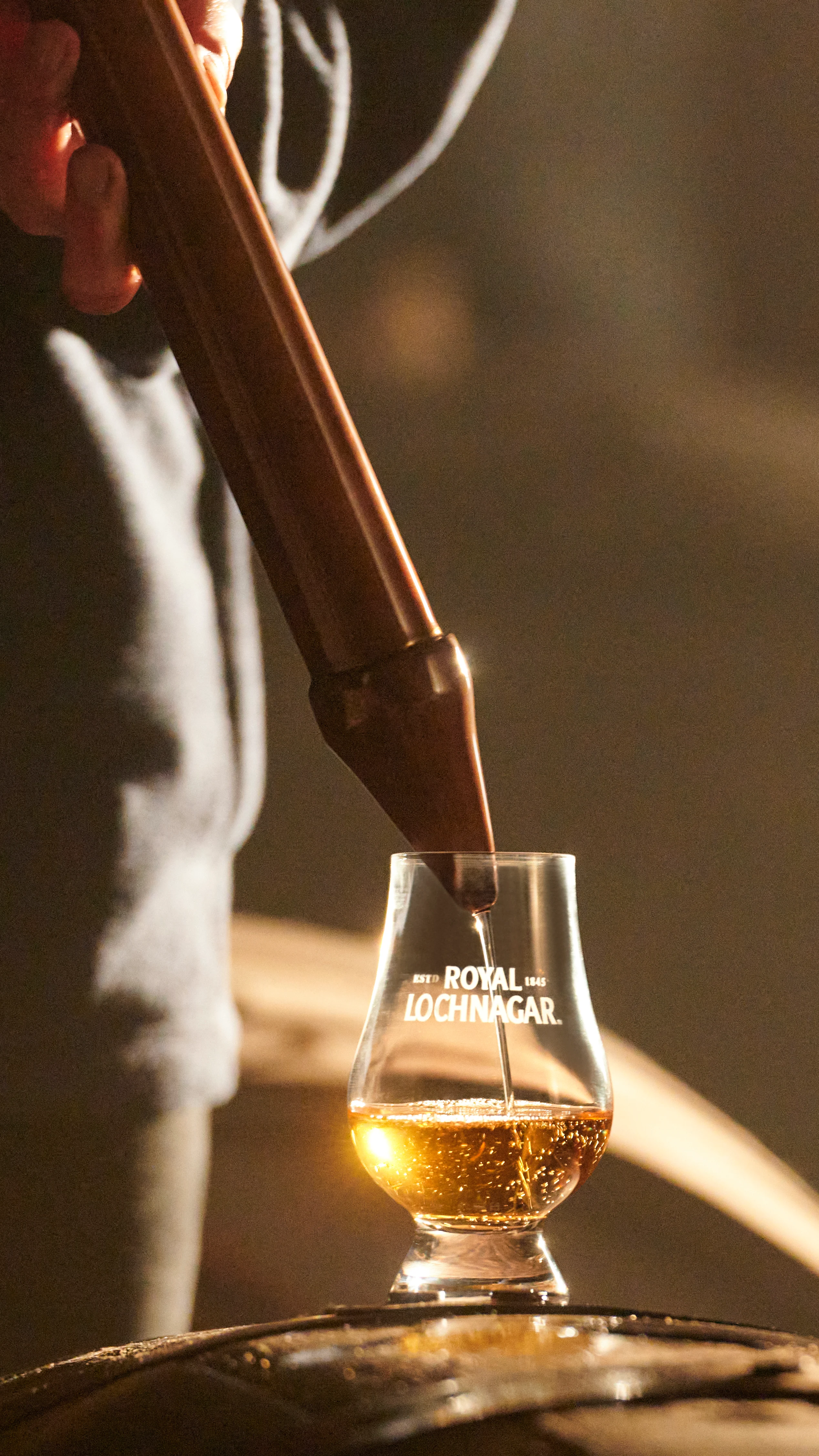Ein kleines Glas mit Whisky steht auf einem Holzfass. Das Glas wird mit einer großen Metallpipette befüllt. Auf dem Glas ist der Schriftzug "Royal Lochnagar" aufgedruckt.