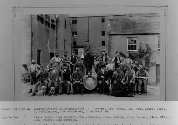 A black and white photo of several men in Victorian dress. The front row of men is sat and in the middle of them is a wooden barrel. Behind the seated men are standing men. Behind the group are buildings.