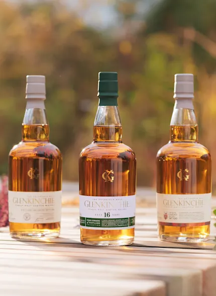 Three bottles of Glenkinchie whisky sit on a row outside on a wooden surface. Surrounding the wooden surface are assorted plants and flowers.