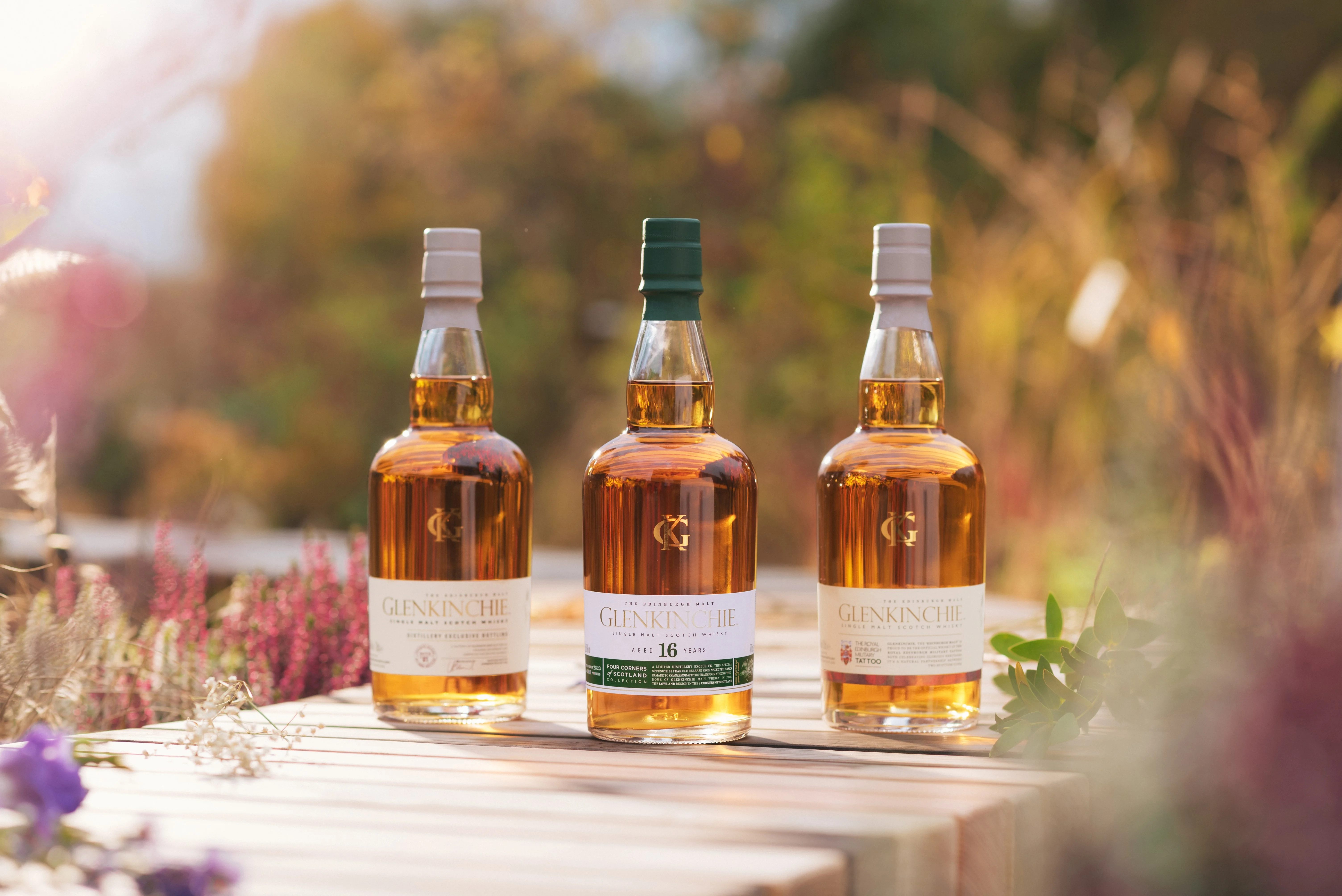 Three bottles of Glenkinchie whisky sit on a row outside on a wooden surface. Surrounding the wooden surface are assorted plants and flowers.