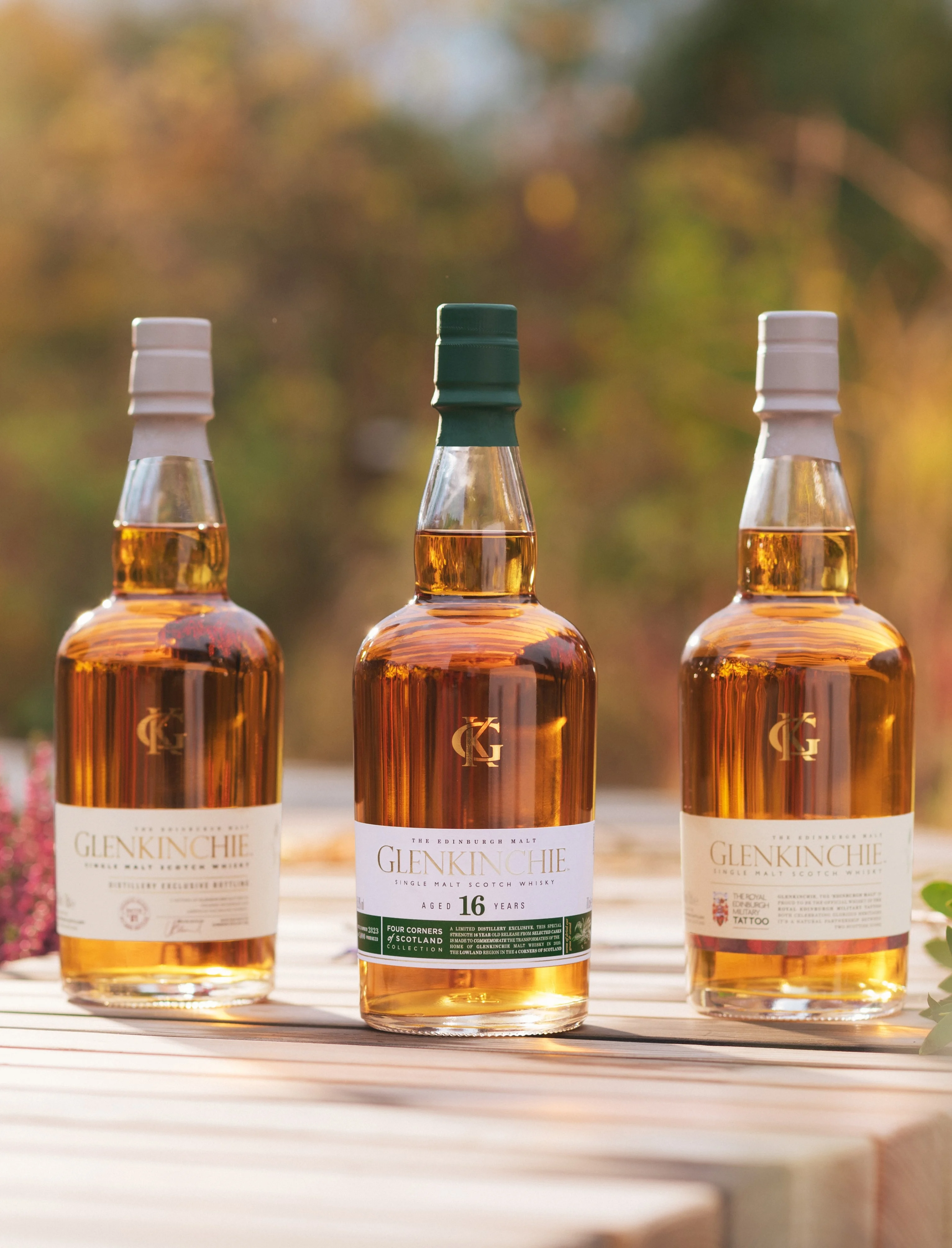 Three bottles of Glenkinchie whisky sit on a row outside on a wooden surface. Surrounding the wooden surface are assorted plants and flowers.