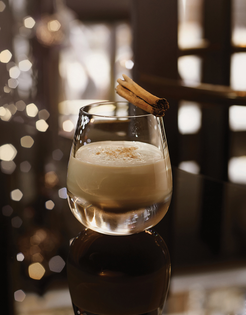 A cocktail with a cinnamon stick and a christmas tree in the background