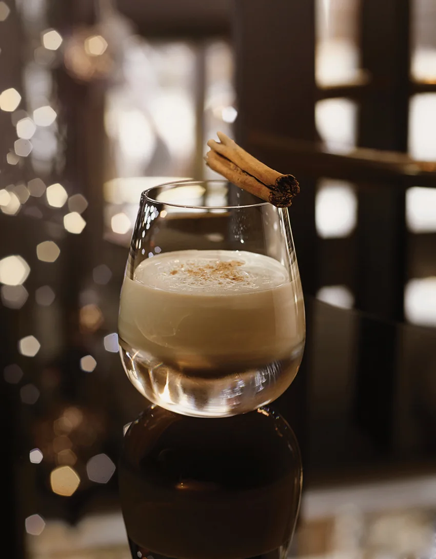 A cocktail with a cinnamon stick and a christmas tree in the background