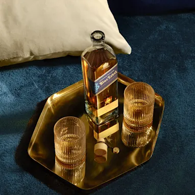 Een fles Johnnie Walker Blue Label staat op een blauw fluwelen oppervlak op een gouden dienblad met twee glazen ijs.