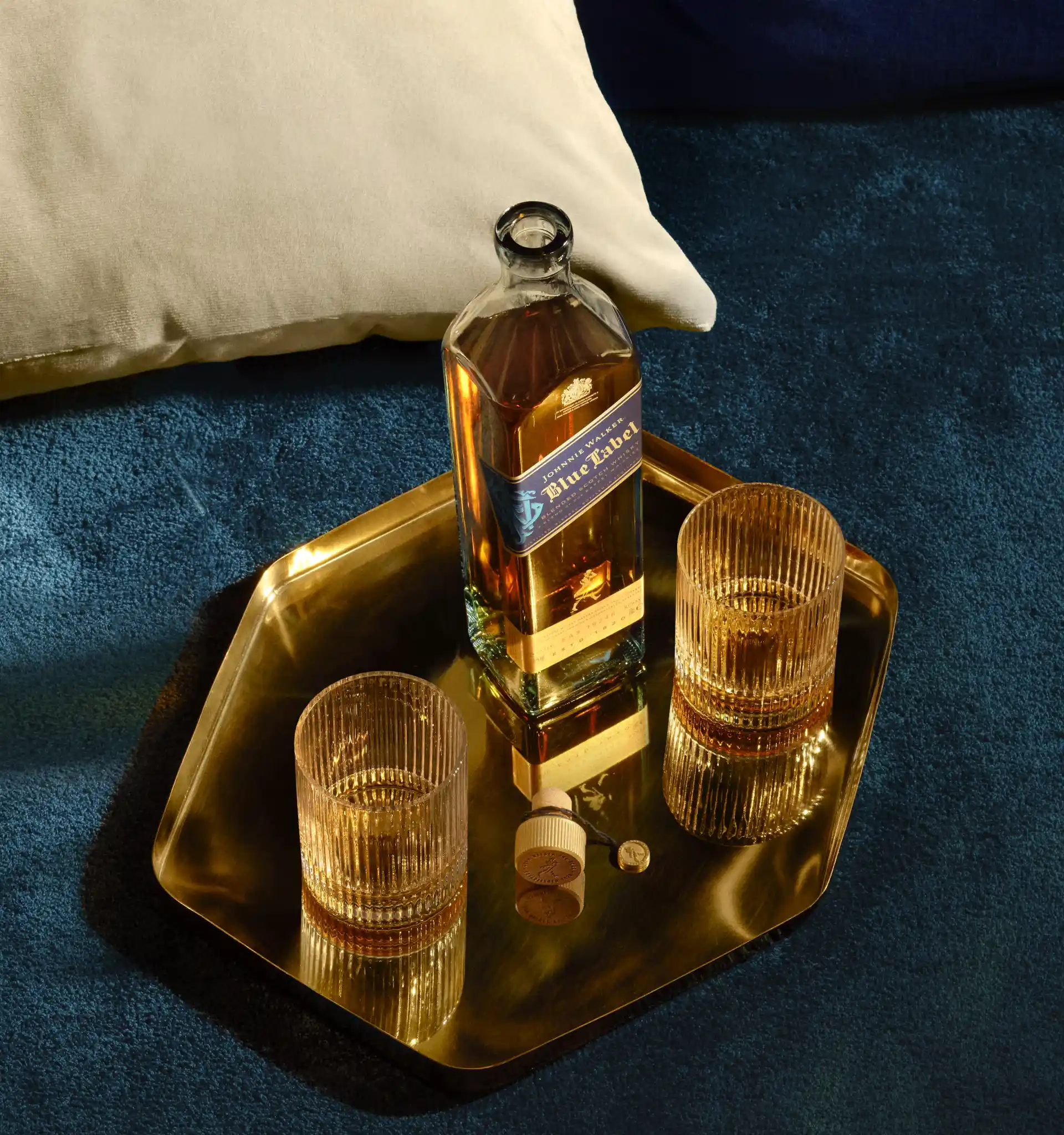 A bottle of Johnnie Walker Blue Label sits on a blue velvet surface on a gold tray with two rocks glasses