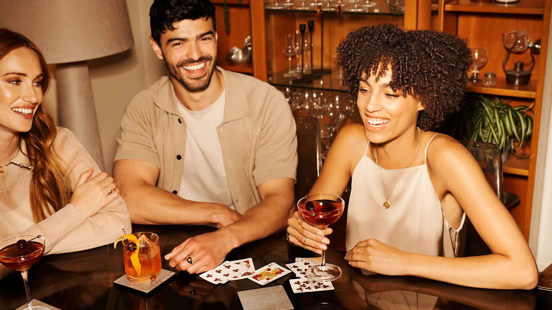 Een man en twee vrouwen zitten aan een tafel. Voor hen staan whiskycocktails en speelkaarten.