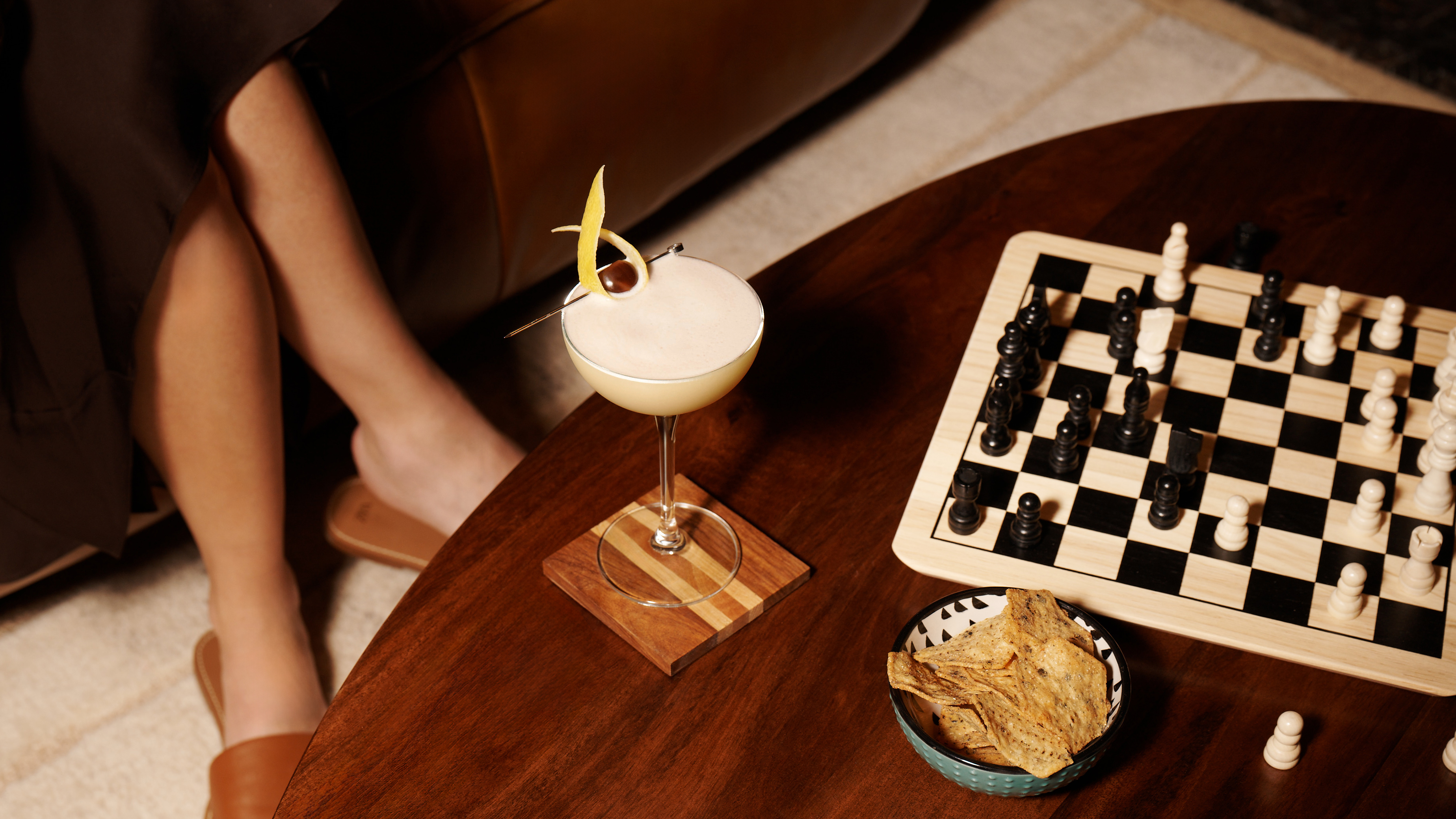 A whisky sour cocktail sits on a coffee table next to a chess board, with a woman sat on a sofa nearby