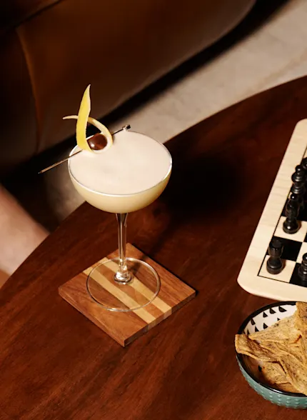 A whisky sour cocktail sits on a coffee table next to a chess board, with a woman sat on a sofa nearby