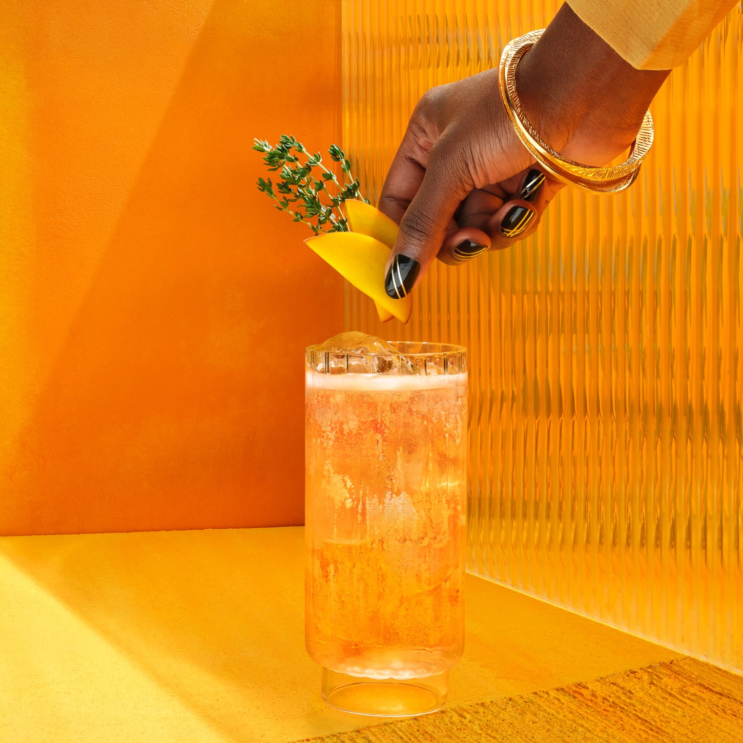  Makkelijke whiskycocktail in een hoog glas met een hand die een garnering toevoegt op een gele achtergrond.