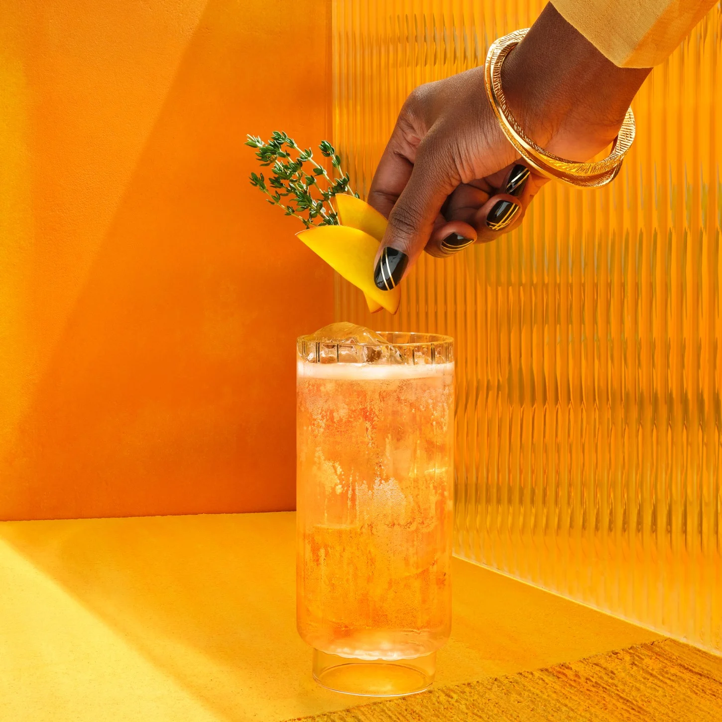  Einfacher Whisky-Cocktail in einem hohen Glas mit einer Hand, die eine Garnierung auf gelbem Hintergrund hinzufügt.