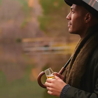 A man stands by a Scottish loch in the autumn. He holds a small tasting glass of whisky in one hand, and the handle of a tartan umbrella in the other hand.