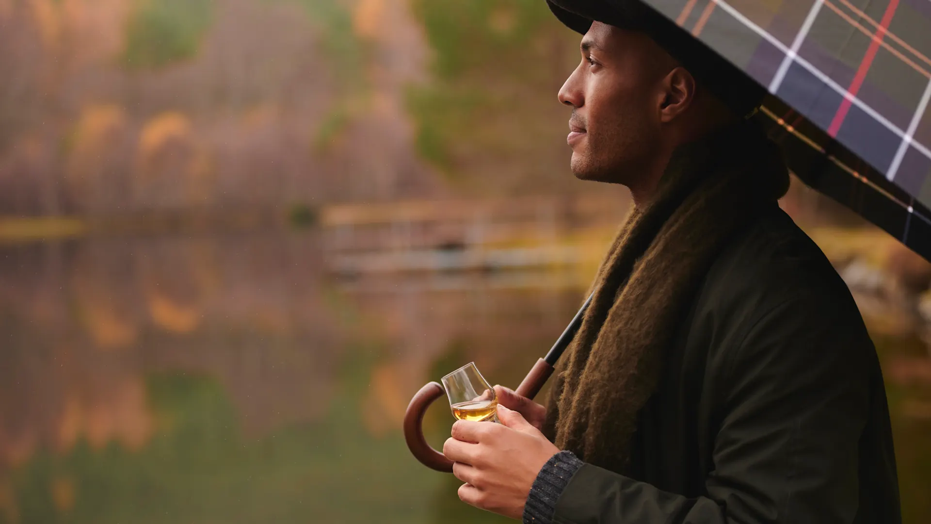 Een man staat bij een Schots loch in de herfst. Hij houdt een klein proefglas whisky in zijn ene hand en het handvat van een ruitjesparaplu in zijn andere hand.