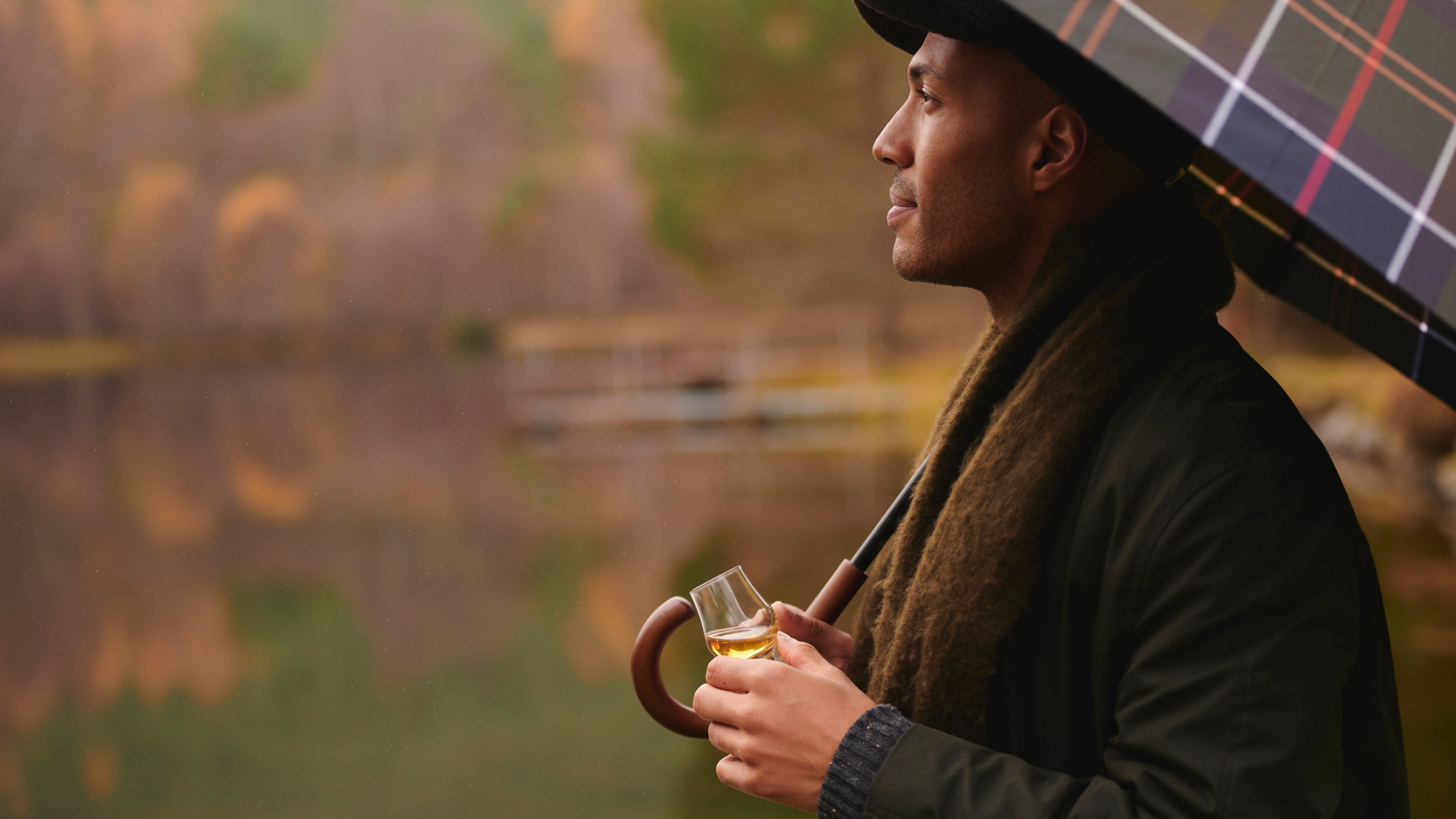 一個男人站在秋天的蘇格蘭湖邊。他一手拿着一小杯威士忌,一手拿着一把格子呢雨伞的伞柄。