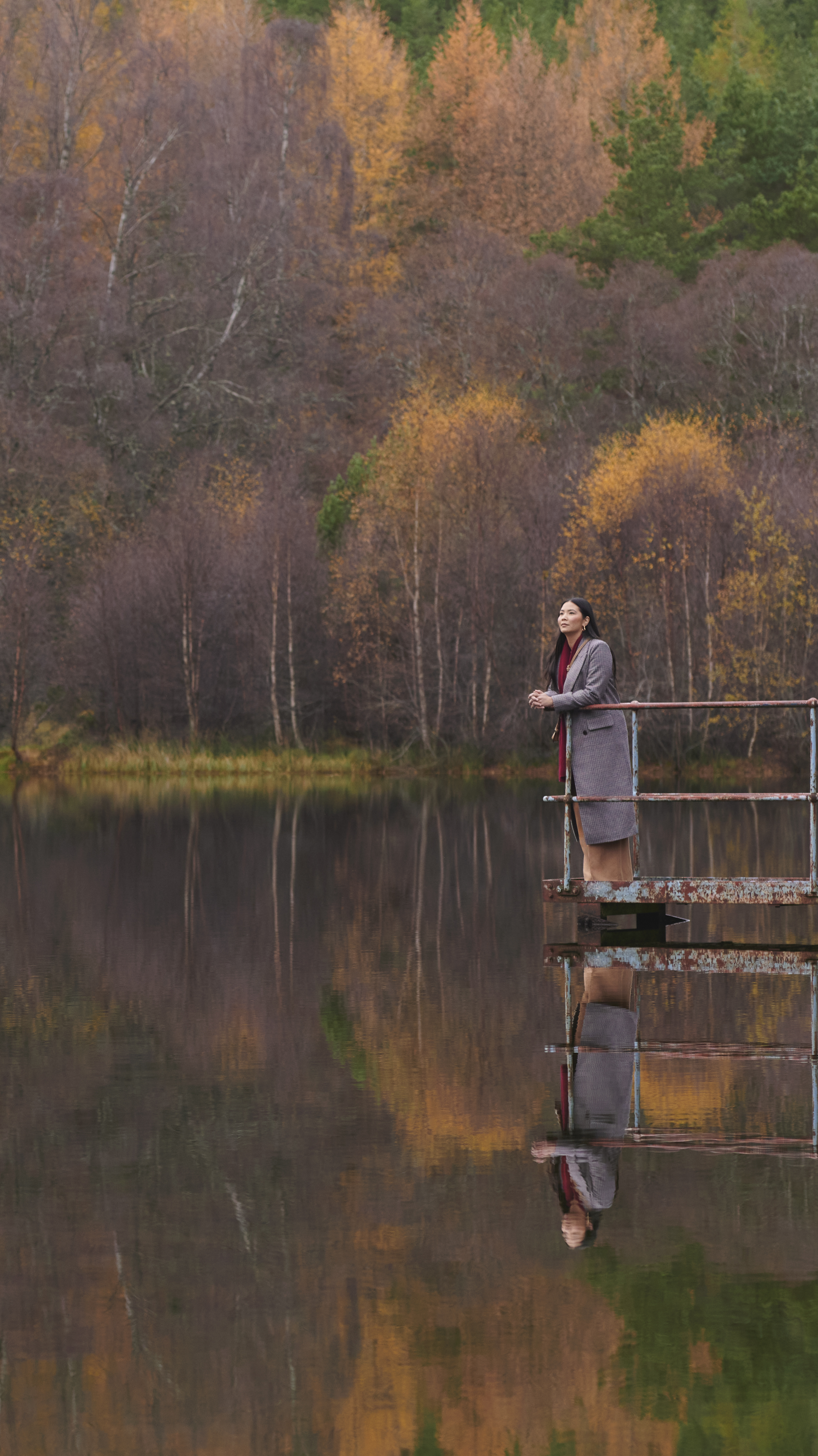 Eine Frau steht auf einem Metallsteg und blickt auf ein schottisches Gewässer. Hinter ihr liegt eine Landschaft mit herbstlichen Bäumen