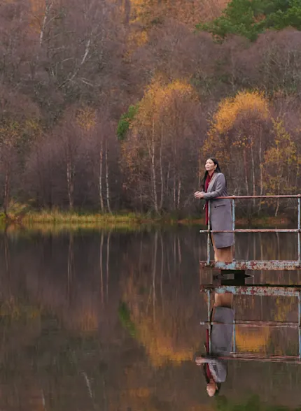 A woman stands on a metal jetty, overlooking a Scottish loch. Behind her is a landscape of autumnal trees