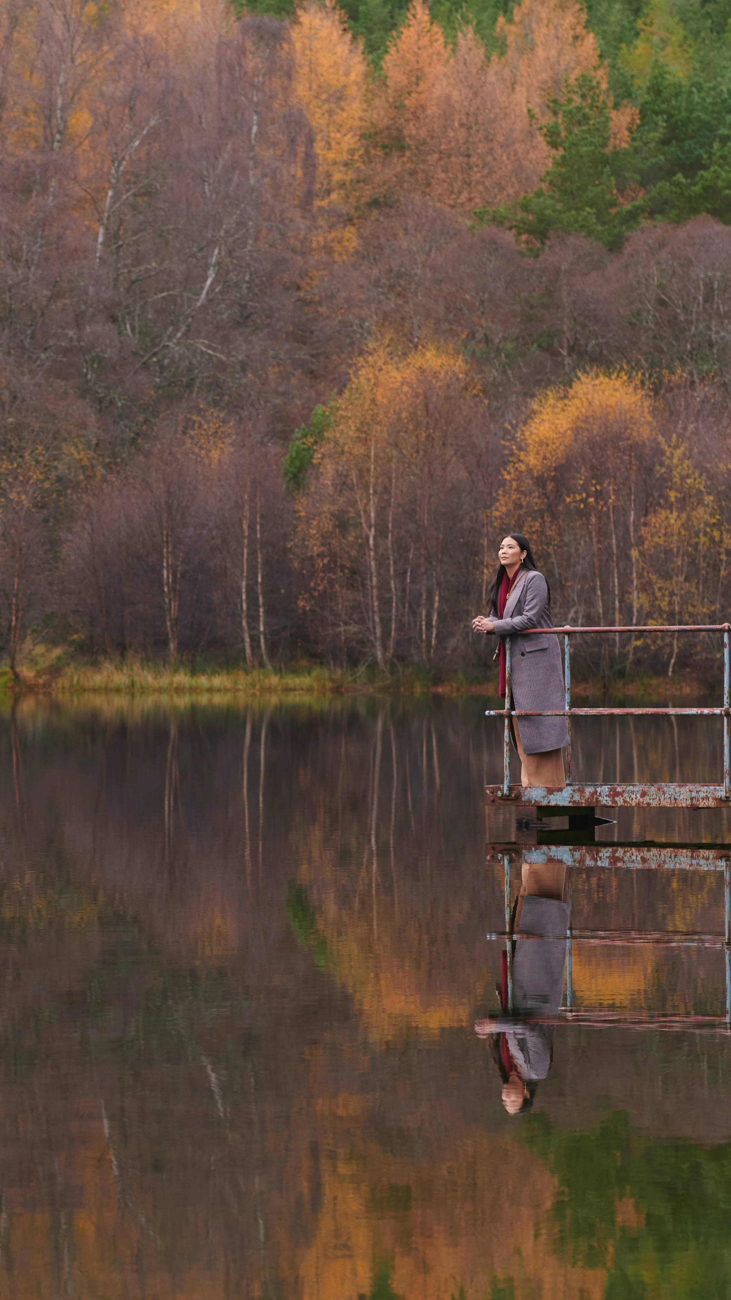 Eine Frau steht auf einem Metallsteg und blickt auf ein schottisches Gewässer. Hinter ihr liegt eine Landschaft mit herbstlichen Bäumen
