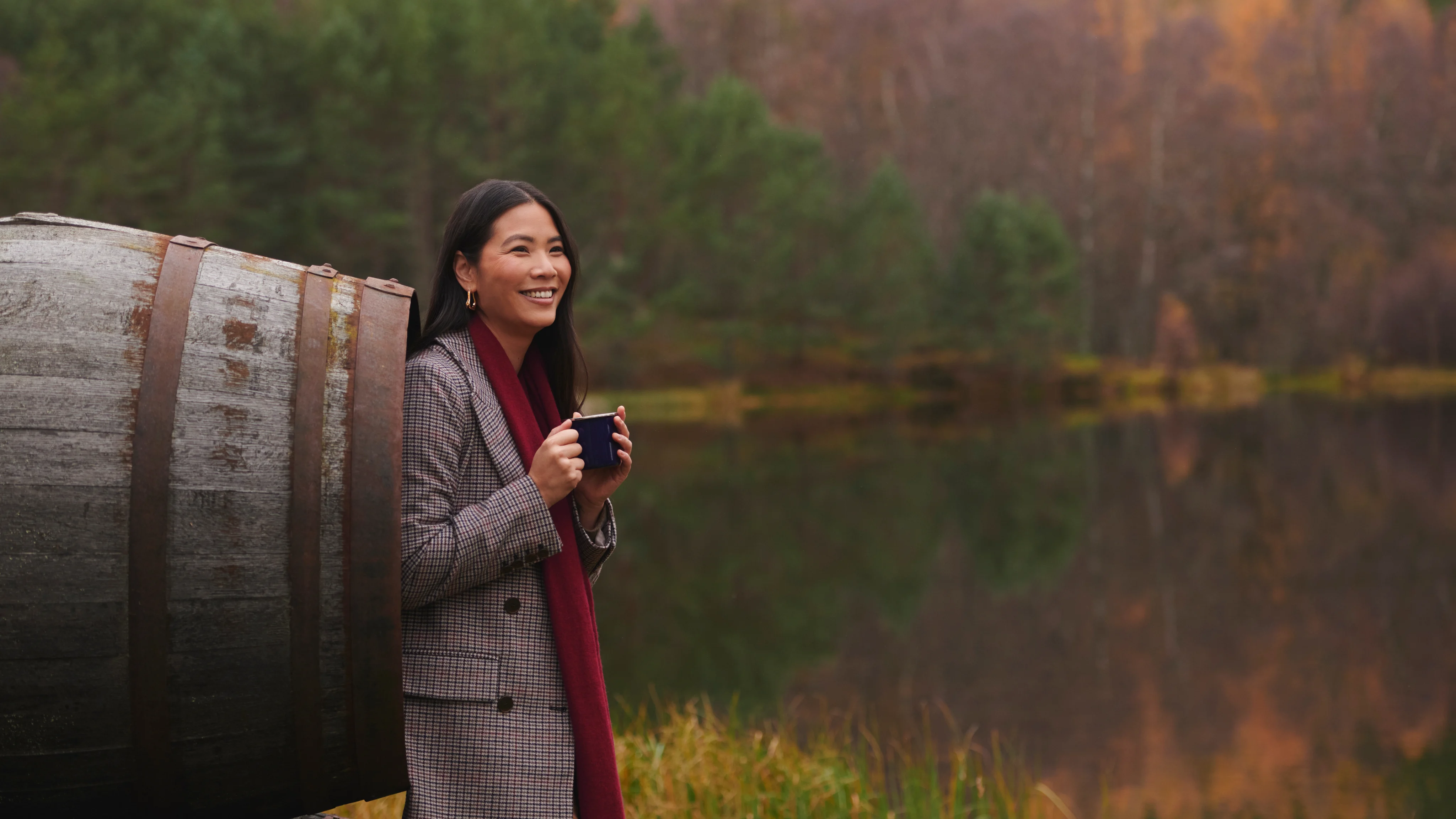A woman leaning against whisky barrel and holding mug, near lake. 