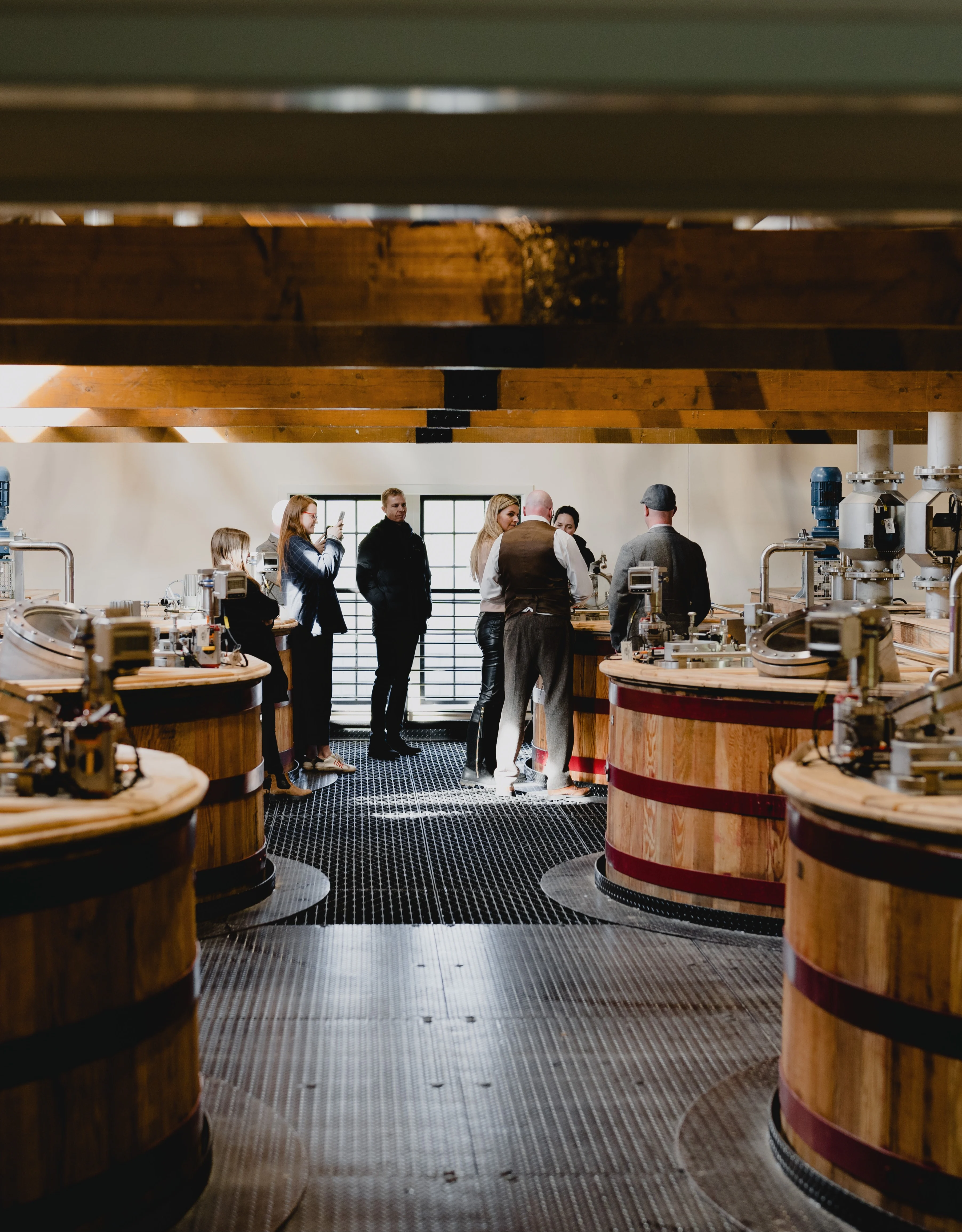 A group of people walk among large barrels while taking a tour inside a distillery.