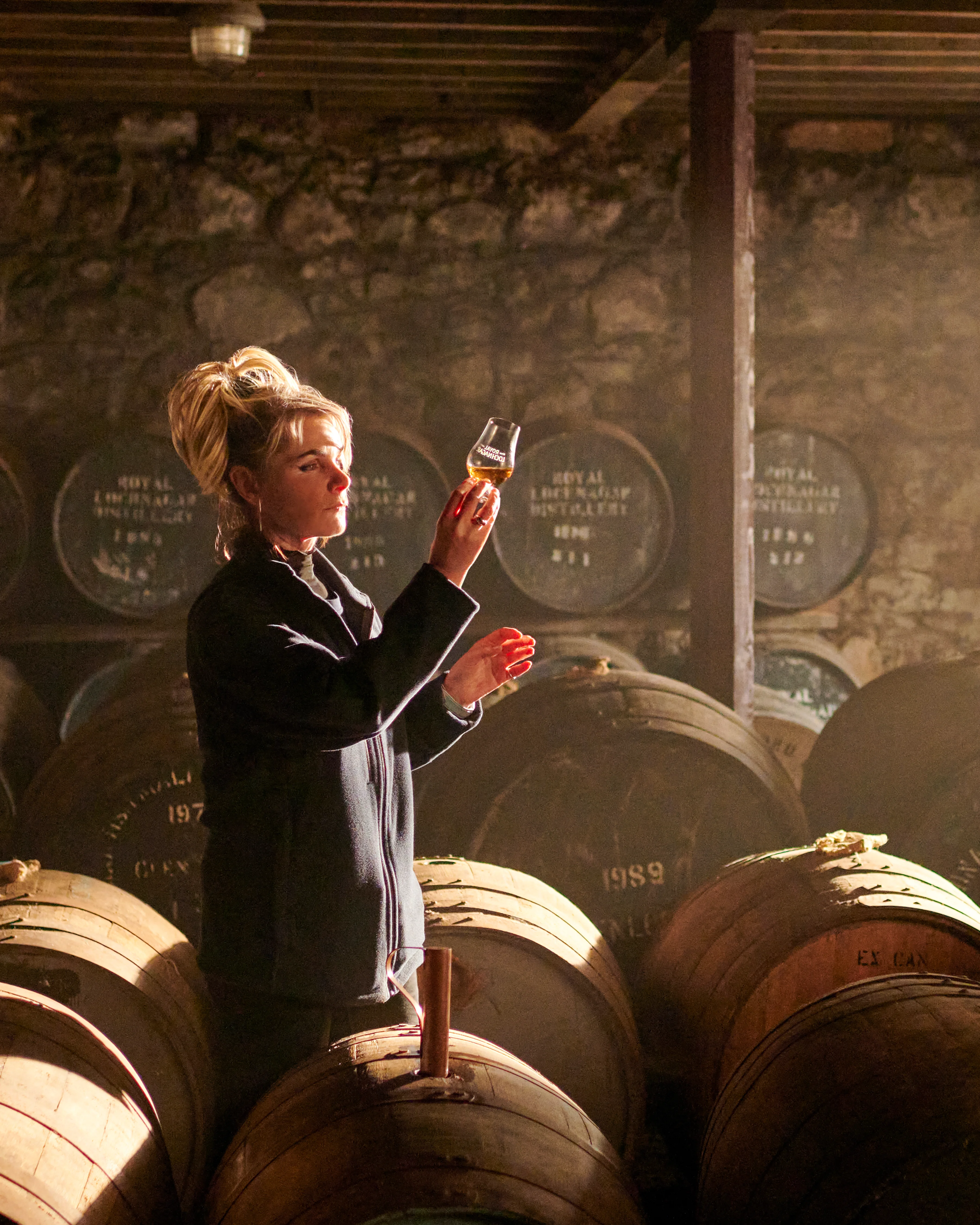A person holding a glass of golden Scotch whisky up to the light in a Scottish Highland distillery setting, with warm amber tones highlighting the whisky's colour