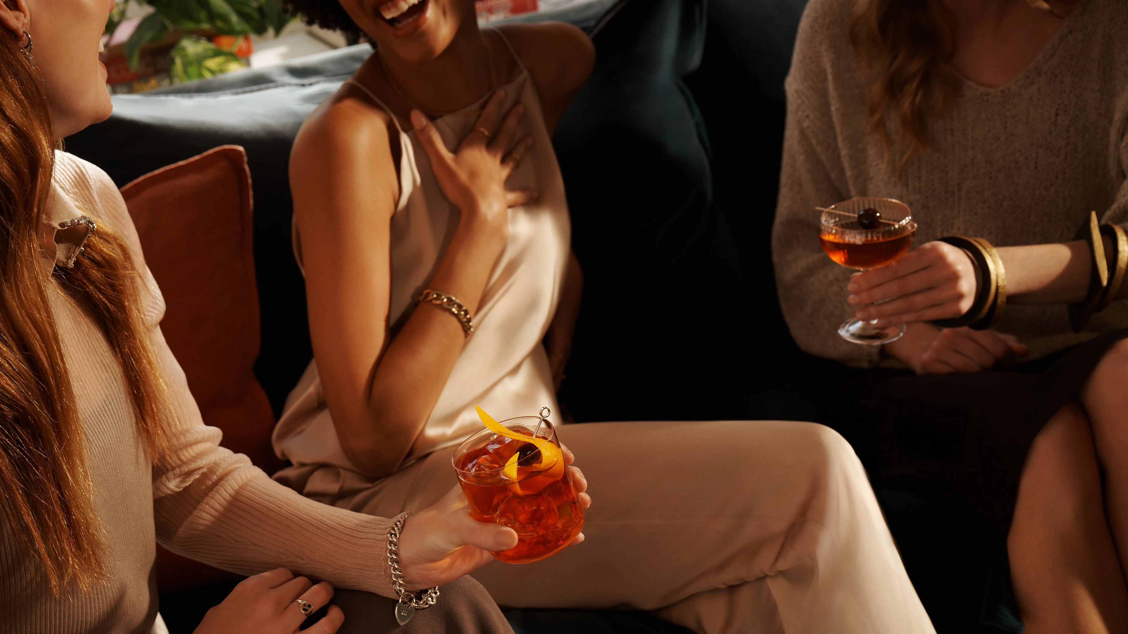 Three women sit on a sofa laughing, one holds an Old Fashioned cocktail and another holds a Manhattan