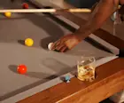 A rocks glass of ice and whisky sits on the edge of a pool table. A man holds a pool cue and repositions a white pool ball.