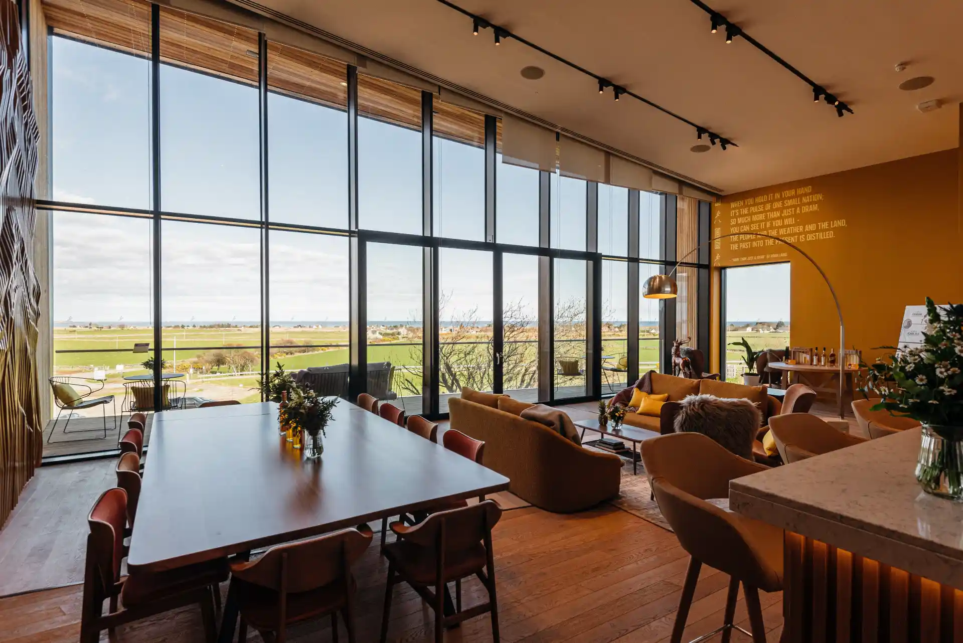 Das Innere des Besucherzentrums von Clynelish mit Holzvertäfelung, Sesseln, Sofas, einer Bar und einem Couchtisch. Eine Wand hat raumhohe Fenster, die auf einen Balkon mit Blick auf grüne Felder führen.