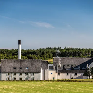 Auchroisk distillery is pictured in front of rolling green hills and a bright blue sky. There is a green lawn in front of the distillery's white building which has a grey roof and a chimney.