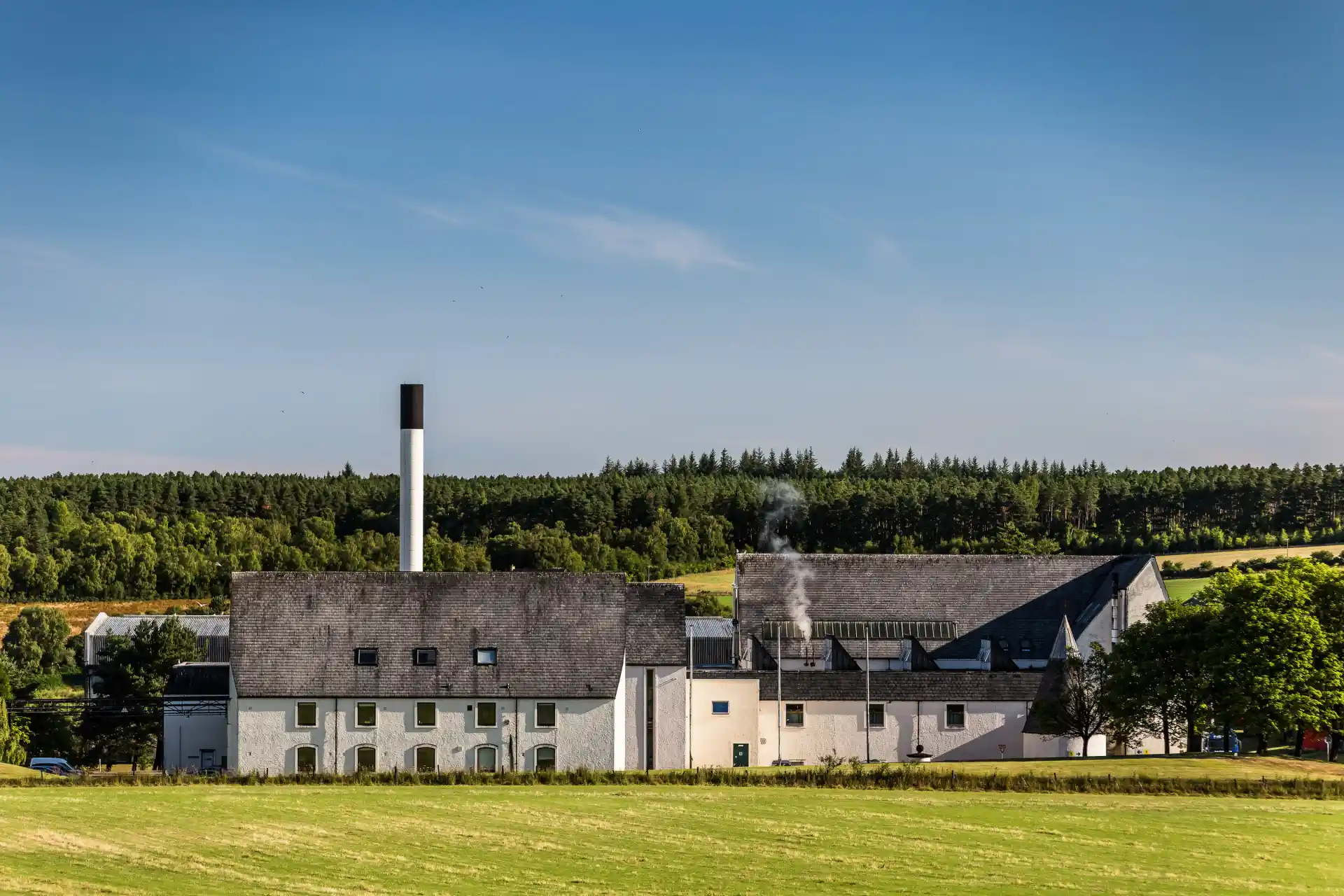 Auchroisk distillery is pictured in front of rolling green hills and a bright blue sky. There is a green lawn in front of the distillery's white building which has a grey roof and a chimney.