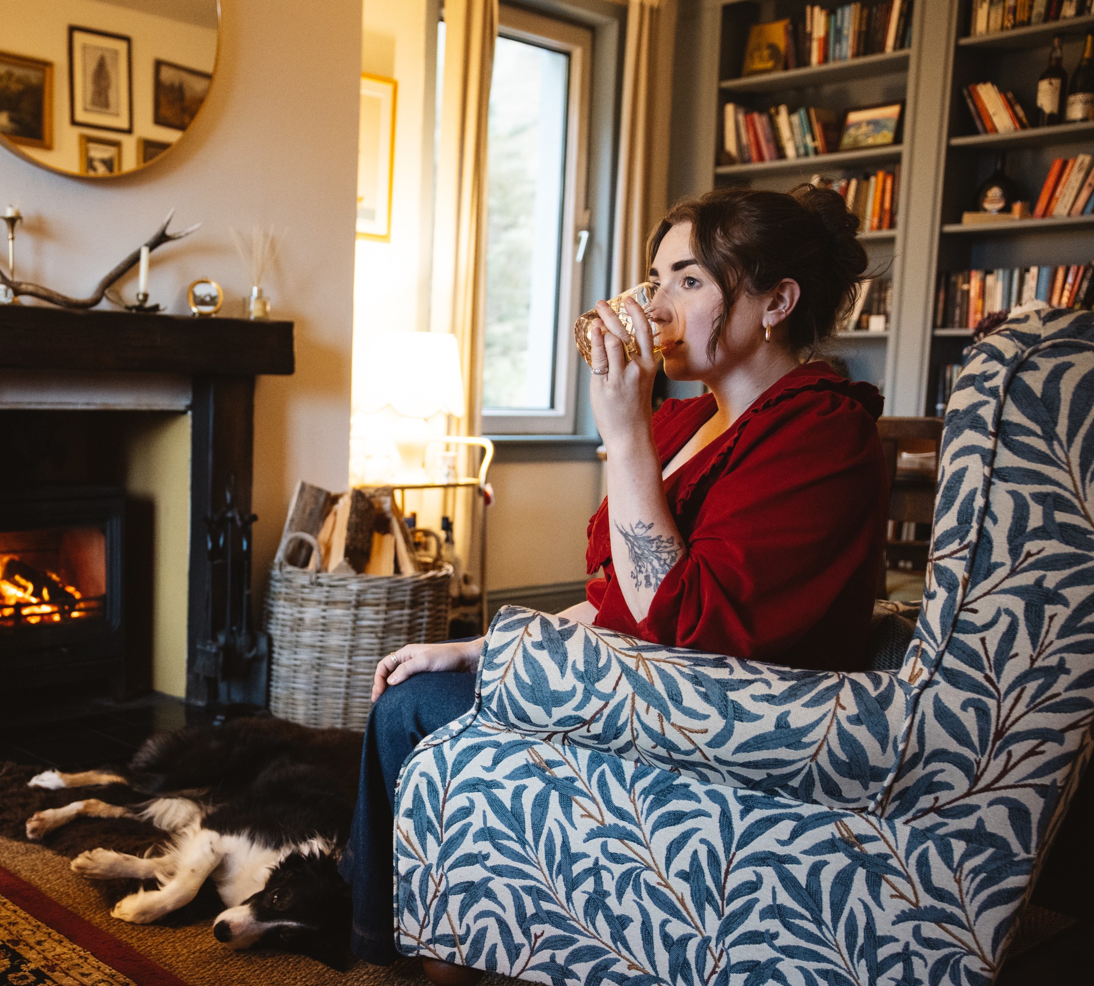 Caroline McQuistin sits in an armchair, sipping whisky from a rocks glass in front of a roaring fire