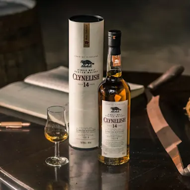 Een klein wit gebouw met een schuin dak in de verte. Op de voorgrond staat een rij gele en groene struiken langs een leeg veld. Rondom het huis zijn velden en een berg in de verte.